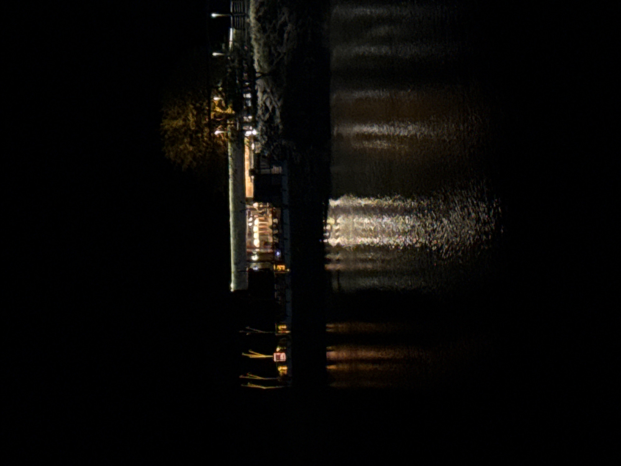 The image depicts a night scene in San Fernando del Valle de Catamarca, Argentina, featuring a body of water reflecting lights from a building or structure situated on the shoreline. The illumination suggests the presence of activity within the building, and lights dot the waters edge. There are light posts and tree shadows on the right. The image is predominantly dark, with light reflections providing the main source of detail.