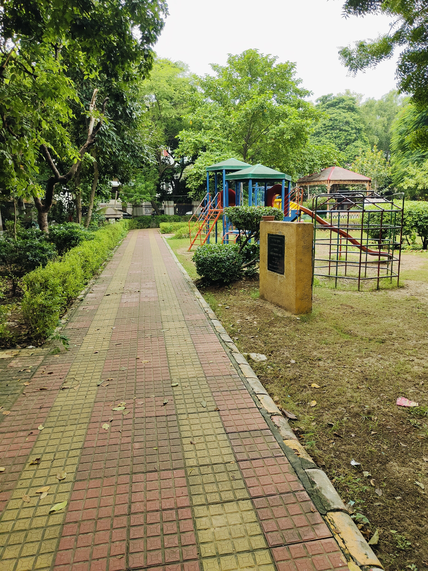 The image depicts a paved walkway in a park in Gurgaon, India. The walkway is constructed with square tiles arranged in a pattern of red and yellow. On the left side of the walkway, there is a neatly trimmed green hedge. On the right side of the walkway, there is a children's playground with slides and climbing structures. A small, brown monument with a plaque is situated near the playground. Beyond the playground, there are trees and greenery, indicating a park setting. The sky is overcast. There are no people visible in the image.