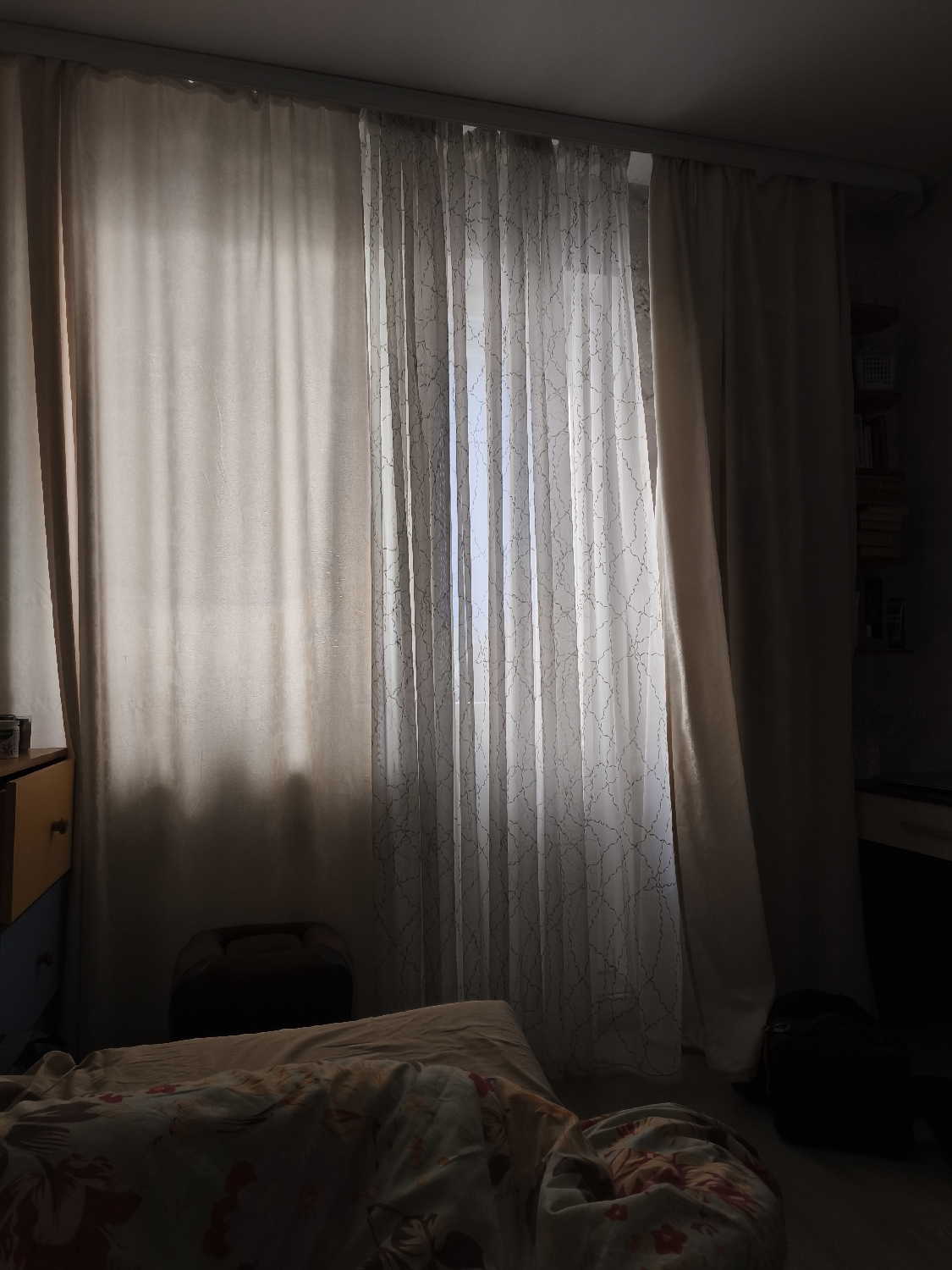 The image depicts an indoor scene, likely a bedroom, characterized by soft, diffused light filtering through curtains. The dominant feature is a large window covered by two layers of curtains: an opaque, light-colored curtain drawn mostly to the left, and a sheer, white or off-white curtain with a delicate, branching pattern covering the entire window. Bright light from outside penetrates the sheer curtain, illuminating its texture and creating a soft glow that casts subtle shadows into the room.
To the left, a portion of a dresser or cabinet with yellow and blue drawers is visible. Below it and extending into the foreground is a bed, partially covered by bedding with a floral or patterned design in shades of red and white. A pillow rests on the bed. In the mid-ground, centered beneath the window, is a dark, low-lying object that might be a stool or a piece of furniture.
To the right of the window, the opaque curtain is drawn, revealing a dark area where shelves holding books or other items can be faintly discerned in the shadows. On the floor to the right, a dark, rounded object, possibly a backpack or a beanbag chair, is visible. The ceiling is plain and light-colored. The overall impression is one of a private, dimly lit space, possibly early morning or late afternoon, designed for comfort and seclusion, with no discernible activity taking place. There is no visible text in the image.