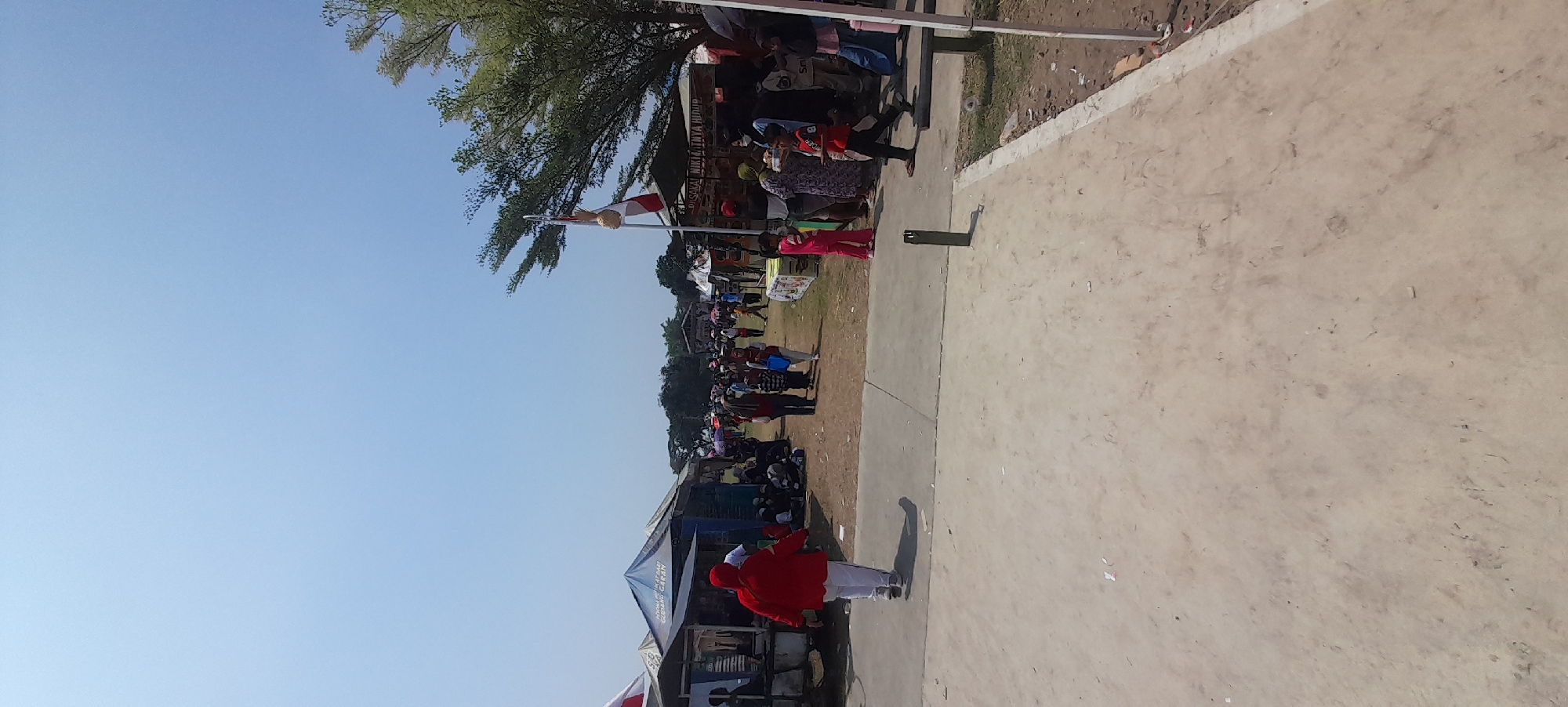 The image depicts a bustling outdoor market scene under a clear blue sky. In the foreground, a person in a red jacket and white pants walks away from the camera on a paved path. The path is bordered by a dirt area where various stalls and tents are set up. Numerous people are browsing the stalls, walking, and socializing. Some tents have signage, one of which reads "PUSAKAI NIKMATNYA HIDUP". A flag with red and white stripes is visible on a pole. Trees with green foliage are scattered throughout the scene, and in the distance, more tents and buildings can be seen. The overall impression is of a lively community event or marketplace. The location is identified as Iser, Indonesia.