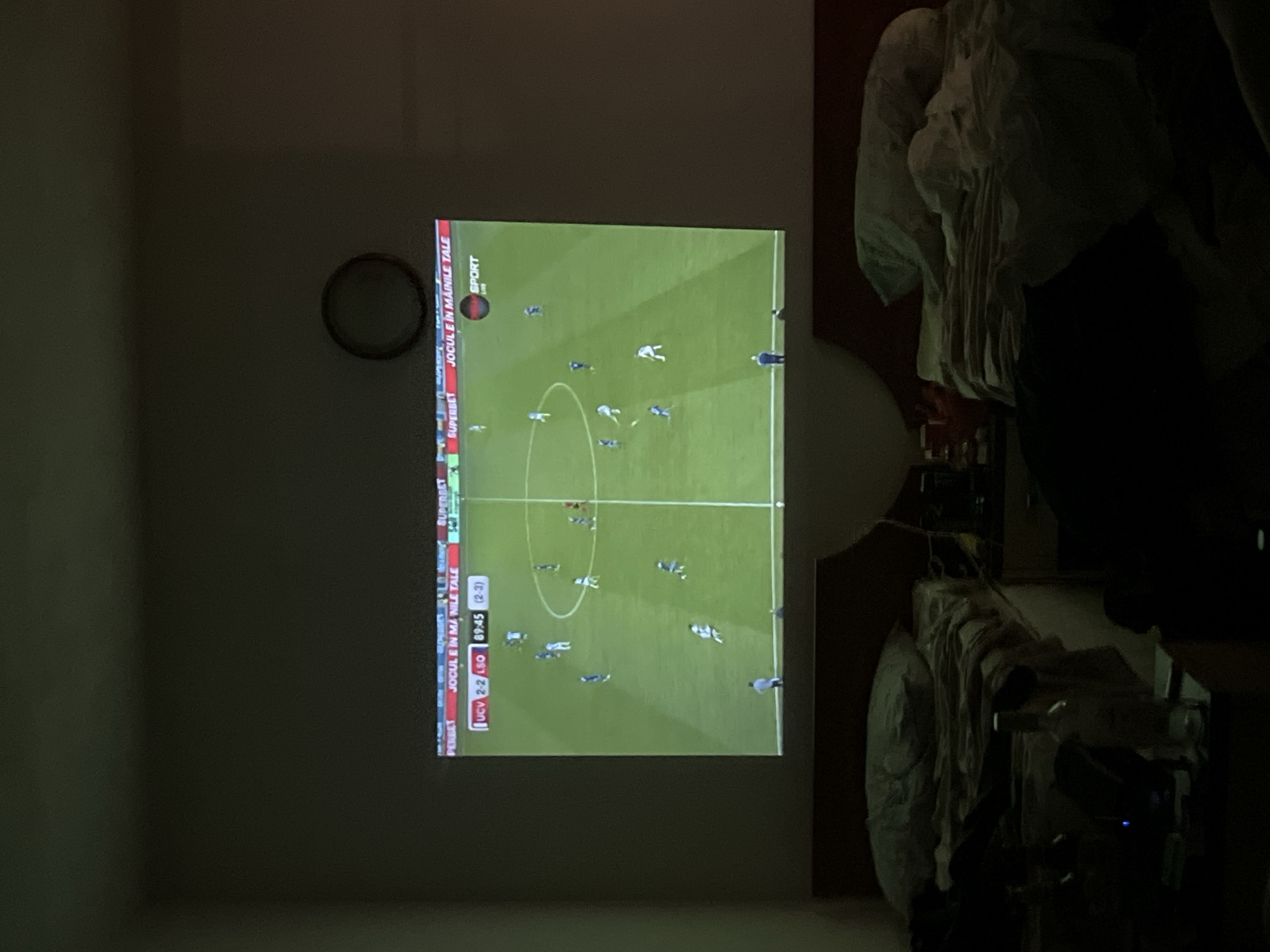 The image captures an indoor scene where a projector displays a soccer match onto a wall. The projected image shows a green soccer field with players in blue and white uniforms. Along the bottom of the projected image, there is text overlay indicating a match score of "UCV 2-2 LSO 89:45 (2-3)". Above the score, text reads "JOCUL E IN MANILE TALE" and "SUPERBET". A circular clock is visible on the wall to the right of the projection. In the foreground, two beds are present, one on the left and one on the right, both with rumpled bedding. The room appears to be dimly lit, suggesting it is nighttime. The overall impression is of a casual viewing experience in a private residence. The city of Obzor, Bulgaria, is not discernible from the visual information provided.