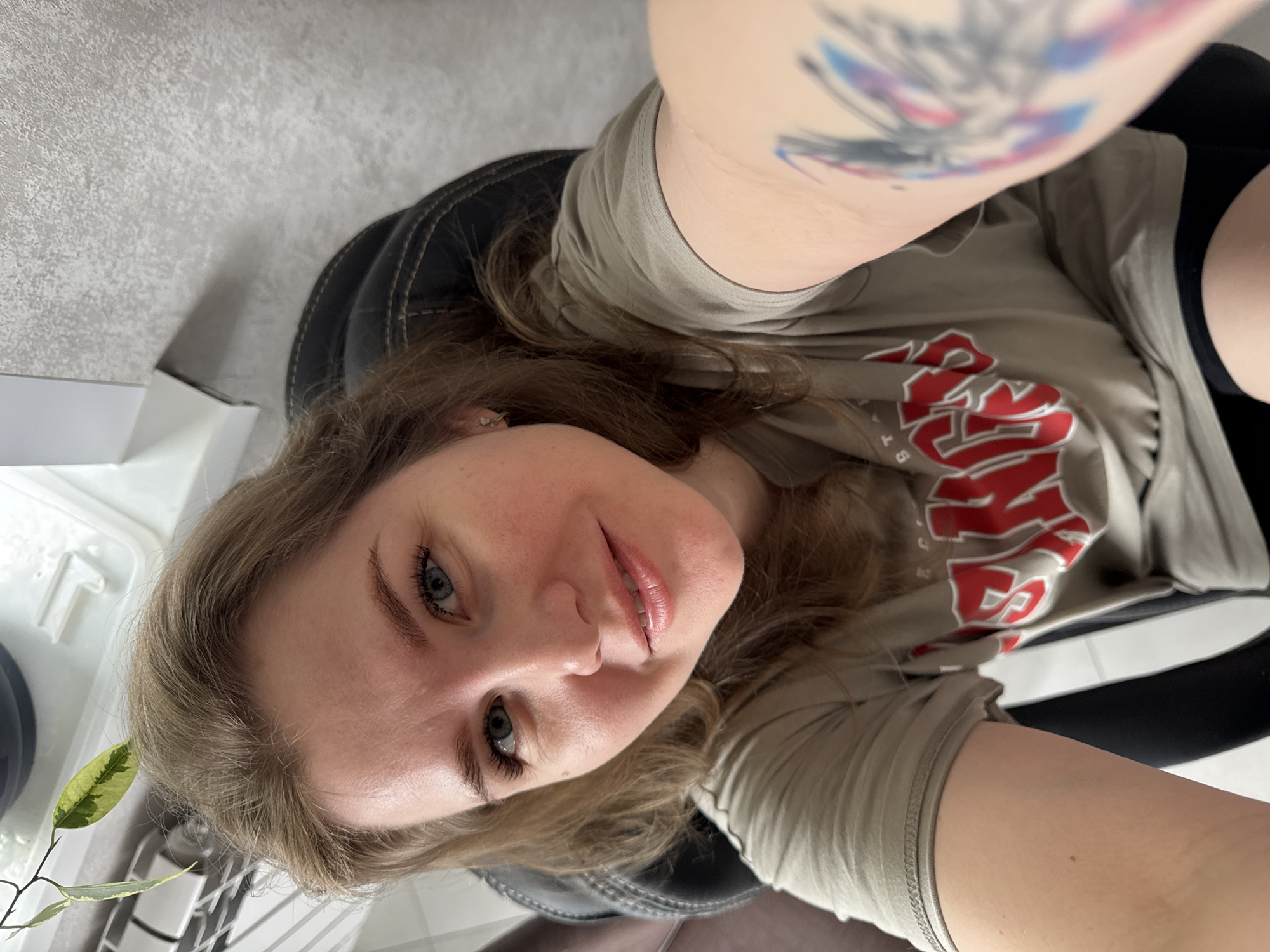 A woman sits in a black office chair, taking a selfie. She has light brown hair and blue eyes. She wears a t-shirt with red lettering. A tattoo is visible on her left arm. The environment appears to be indoors, with a glimpse of a white surface and plant visible in the lower left corner. There is no definitive information to establish the location in Gornyi, Russia.