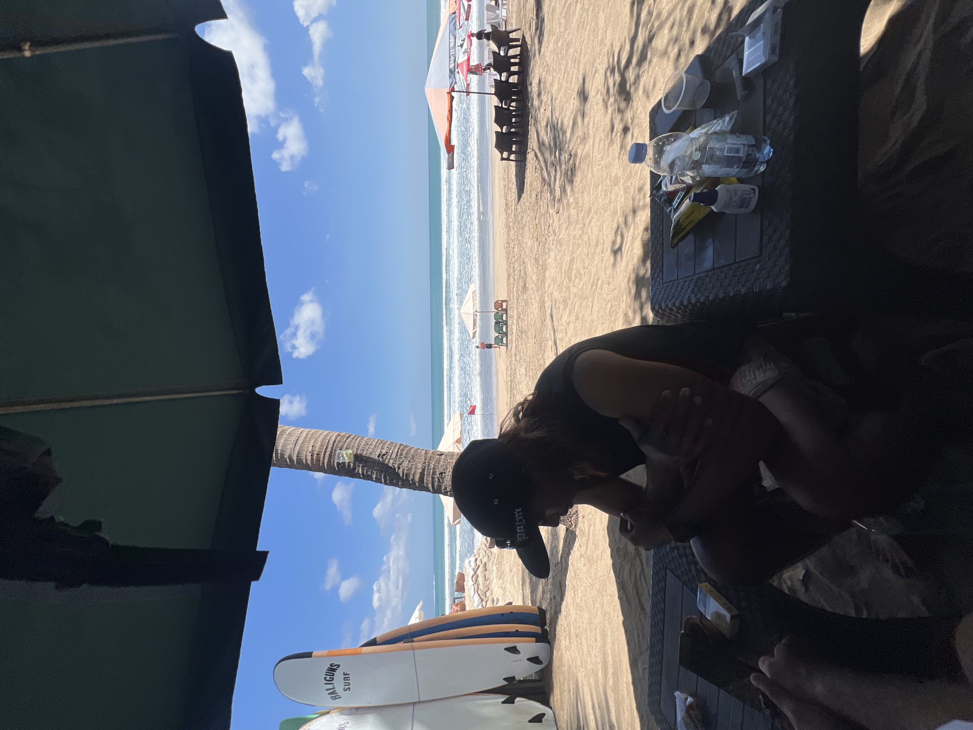 The image depicts a beachfront scene in Kuta, Indonesia. In the foreground, a person wearing a black baseball cap and a dark t-shirt is seated at a table. The table is cluttered with various items including a water bottle, a small white bottle, a packet of cigarettes, and other unidentifiable objects. The person appears to be looking down, with their arms crossed. To the left of the foreground subject, a stack of surfboards is visible, with the branding "BALIGUNS SURF" clearly legible on one of them. In the mid-ground, the beach is populated with lounge chairs and umbrellas, with several people scattered around. The ocean stretches out to the horizon under a clear blue sky with scattered clouds. A prominent palm tree stands to the right of the surfboards. The overall setting suggests a relaxed atmosphere at a beach resort.
