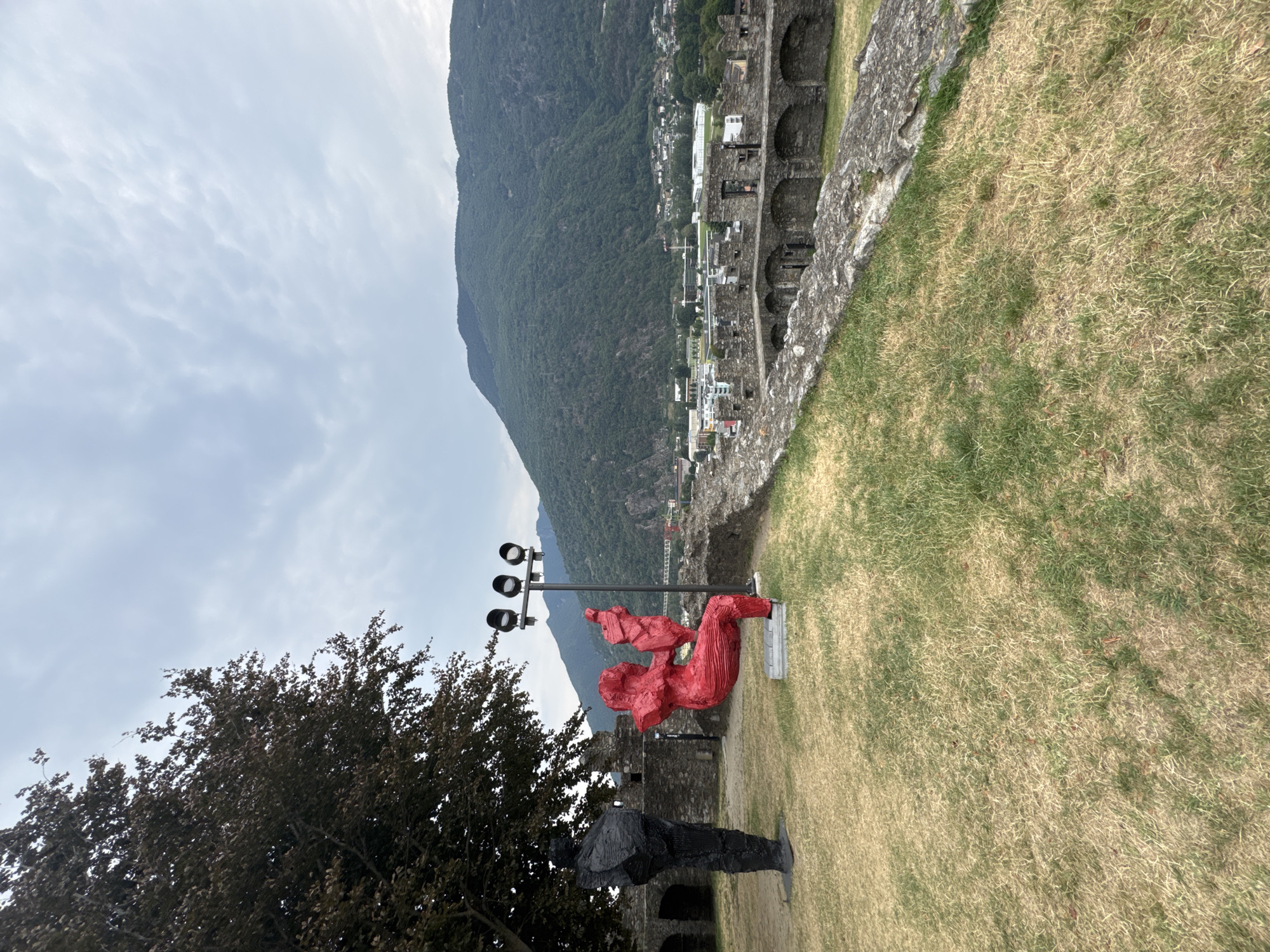 An exterior, medium shot captures a section of the Castel Grande in Bellinzona, Switzerland, overlooking a town and a mountain range under a cloudy sky. In the foreground, two outdoor sculptures are positioned on a grassy area. To the left, a tall, black, abstract human figure stands. To the right of the black figure, a bright red abstract human sculpture is seated, appearing to be on a swing. The red sculpture is holding a smaller, abstract object. Behind the sculptures, the stone walls and arched structures of the Castel Grande are visible. Beyond the castle walls, the town of Bellinzona spreads out, with buildings and infrastructure extending towards a large, green, tree-covered mountain that dominates the background. A light pole with three spotlights stands between the two sculptures. The grass in the foreground is dry and patchy in some areas. The overall lighting suggests it is daytime.