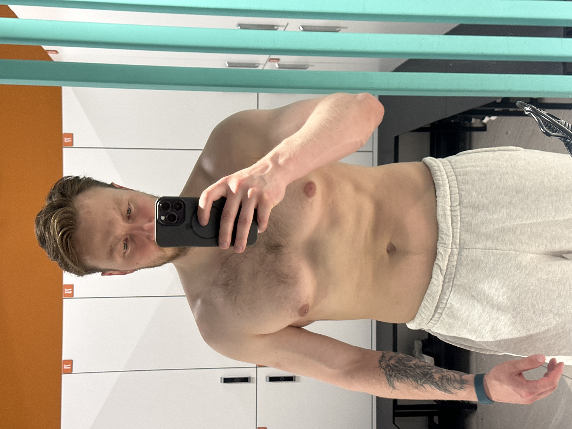 A light-skinned man is taking a selfie in a mirrored room. He is shirtless and wearing gray sweatpants. A black phone is held in his right hand. The background reveals lockers with orange and white doors, a teal-colored horizontal slatted structure, and what appears to be a gym setup. The man has short blonde hair, and a visible tattoo on his left forearm. The location is not discernible as Sochi, Russia, from the provided information.