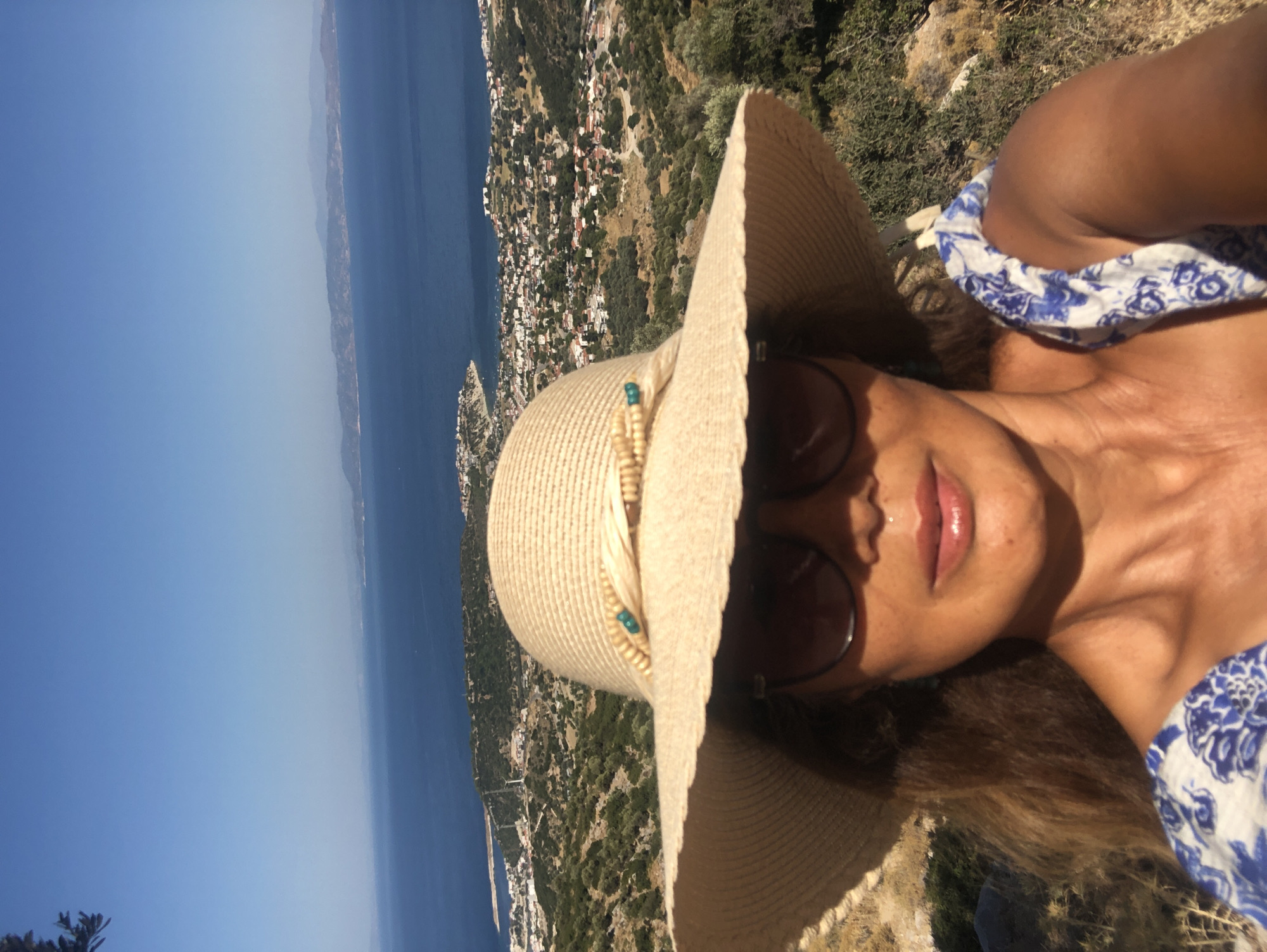 A woman is captured in a selfie, wearing a wide-brimmed straw hat and sunglasses, with a blue and white patterned top visible. She is standing outdoors, with a panoramic view of the Aegean Sea and a coastal town. The landscape features hills covered in sparse vegetation and olive trees, leading down to the town with white buildings and red roofs. In the distance, mountains rise beyond the sea, under a clear blue sky. The location is identified as the city of Karaburun, Turkey.