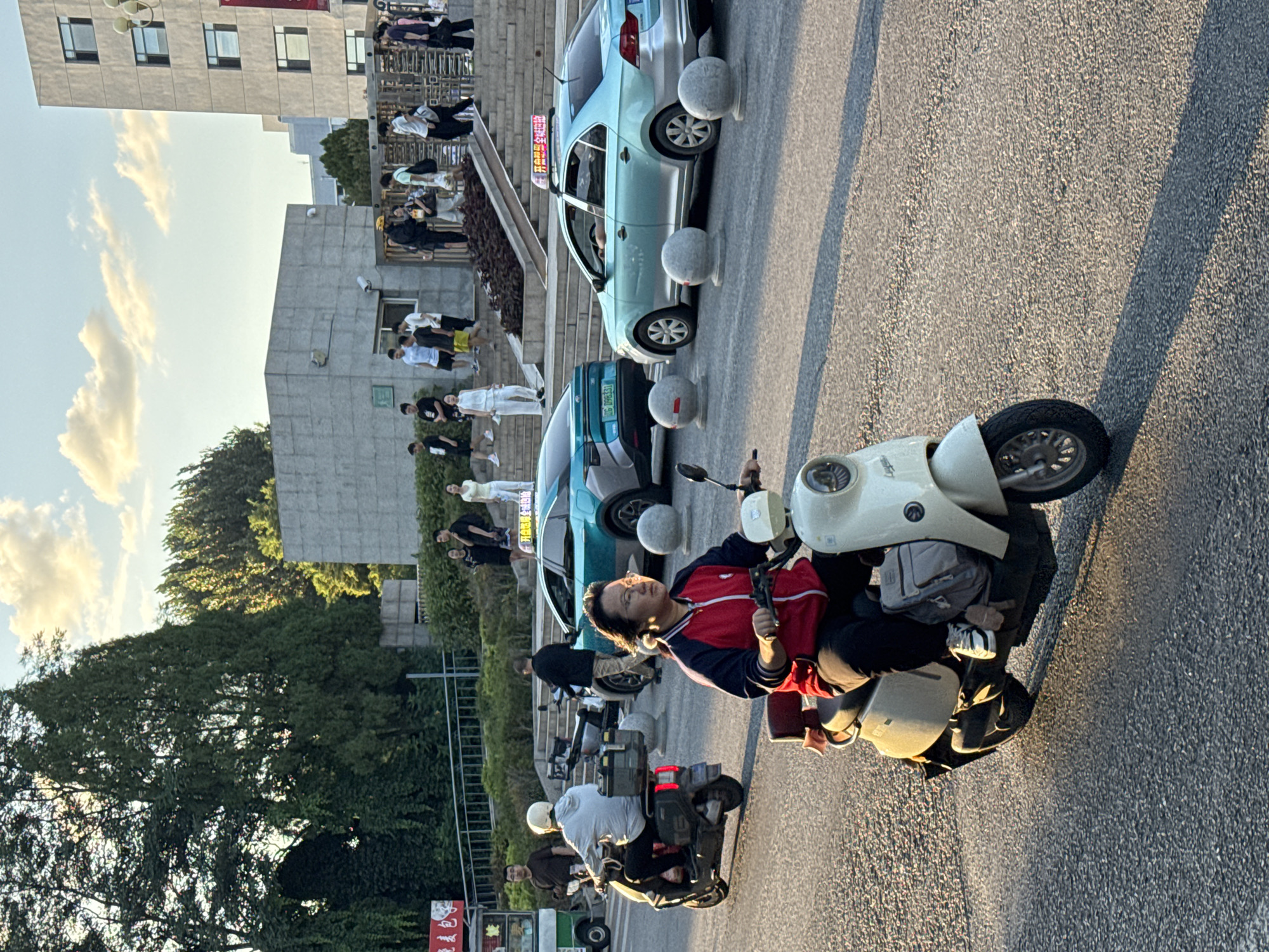 The image depicts a street scene in Zhongtun, China, during the late afternoon or early evening, as indicated by the warm, long shadows. In the foreground, a person wearing a red and blue jacket, dark pants, and white sneakers is riding a light-colored electric scooter. The scooter has a cream-colored body and a brown seat, with a gray bag strapped to the rear. The person is looking forward, with glasses on and their hair tied back.
Further back, on the left side of the frame, another individual on a black electric scooter is wearing a white helmet and a light blue hooded jacket. A food cart with red and white signage is partially visible behind them.
In the middle ground, two light blue cars are parked or moving on the street. The car on the right has a taxi sign on its roof with Chinese characters. Several large, rounded concrete bollards are placed along the roadway.
In the background, a modern building with multiple windows stands tall. Steps lead up to an entrance area where a group of people are visible. Some are walking up the stairs, while others are standing and conversing. The environment includes trees and greenery, suggesting a well-maintained urban area. The sky is partly cloudy, with soft sunlight filtering through.
