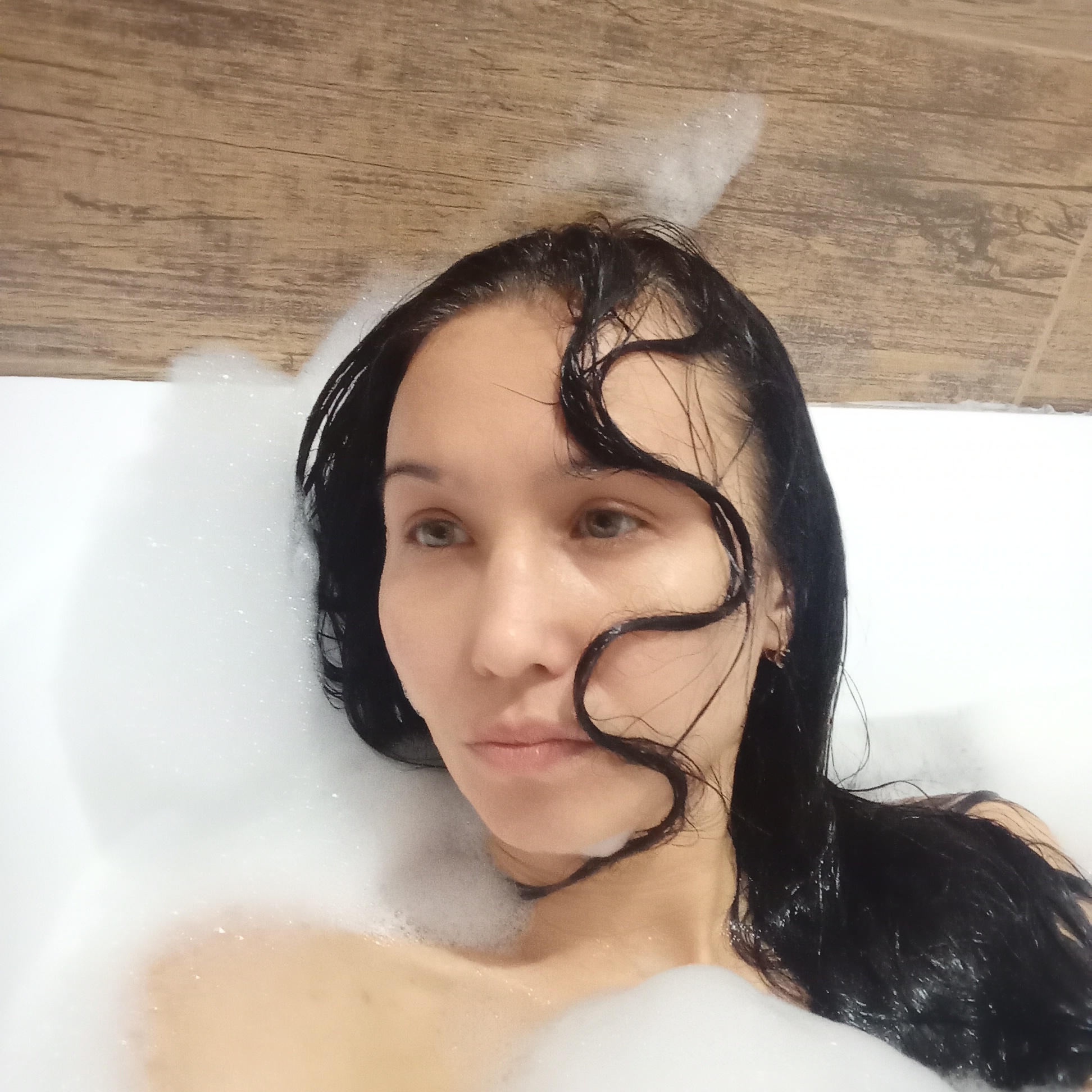 A woman with wet, dark hair is submerged in a white bathtub filled with soapy bubbles. The woman's face is visible and she is looking to her right. A section of wall with a wood-grain pattern is visible at the top of the frame. There is no evidence that this image was taken in Pervomaysky, Russia.