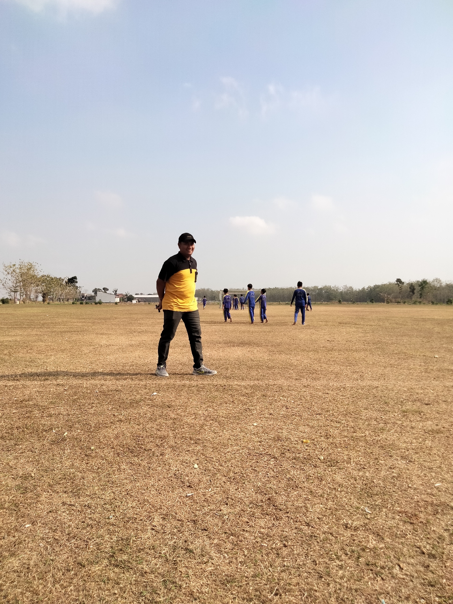 A man in a yellow and black shirt and black pants stands on a dry, grassy field, smiling and looking to his right. He is wearing a black baseball cap and grey sneakers. In the background, several individuals in blue uniforms are walking across the field. A soccer goal is visible behind them, and trees line the horizon. The sky is clear with a few wispy clouds. The scene appears to be a sports field in Pucakwangi, Indonesia.