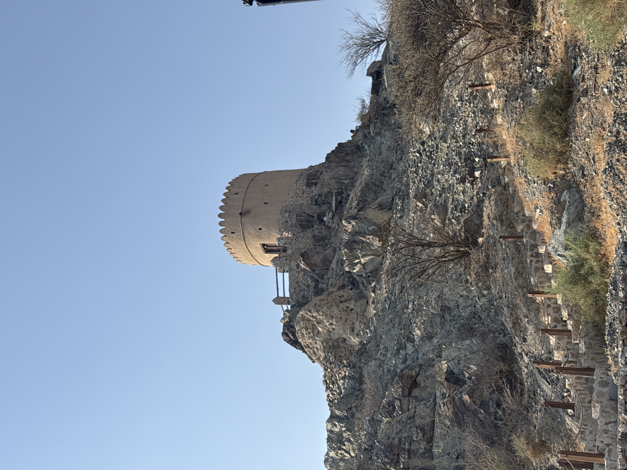 A historic Hatta Fort, a circular tower made of mudbrick and stone, sits atop a rocky hill in Hatta, United Arab Emirates. The fort has a crenellated parapet and a single entrance. A wooden fence surrounds the fort's viewpoint. A stone staircase, lined with wooden posts and rope railings, winds its way up the rocky slope towards the fort. Sparse, dry vegetation, including small trees and shrubs, dots the arid landscape. The sky is a clear, bright blue.