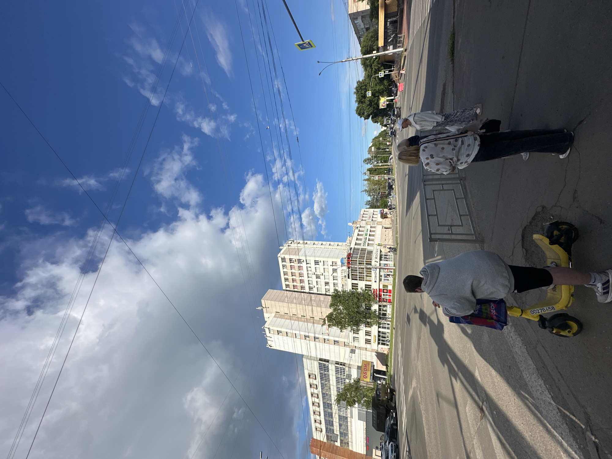 The image depicts an outdoor scene in Rubtsovo, Russia. The primary subjects are three people, two women and one man, interacting on a street. One woman is standing and looking away from the camera, wearing a patterned shirt and dark pants. Another woman, with blonde hair, is also standing, wearing a white patterned shirt and dark pants, and has a bag slung over her shoulder. The man is seated on a yellow electric scooter, wearing a grey hooded sweatshirt and black shorts, looking down at something on the scooter.
The setting appears to be a city street with a sidewalk and a road. In the background, several tall buildings line the street, some of which have visible signage. Green trees are also present, suggesting a relatively urban environment. The sky is a vibrant blue with scattered white clouds, indicating fair weather. Power lines are prominently visible, crisscrossing the sky.
The activity appears to be casual. The people are on a street, with one individual on a scooter, suggesting they might be commuting, sightseeing, or simply taking a break. The time of day is likely afternoon, given the bright sunlight and long shadows cast on the ground. There's no indication of a specific event or strong emotions.
Visible text includes what appears to be the number "00278458" on the yellow scooter. Other text is present on building signage and a street sign, but it is too small or blurry to be legible in detail. The overall impression is a snapshot of everyday life on a street in a Russian city.