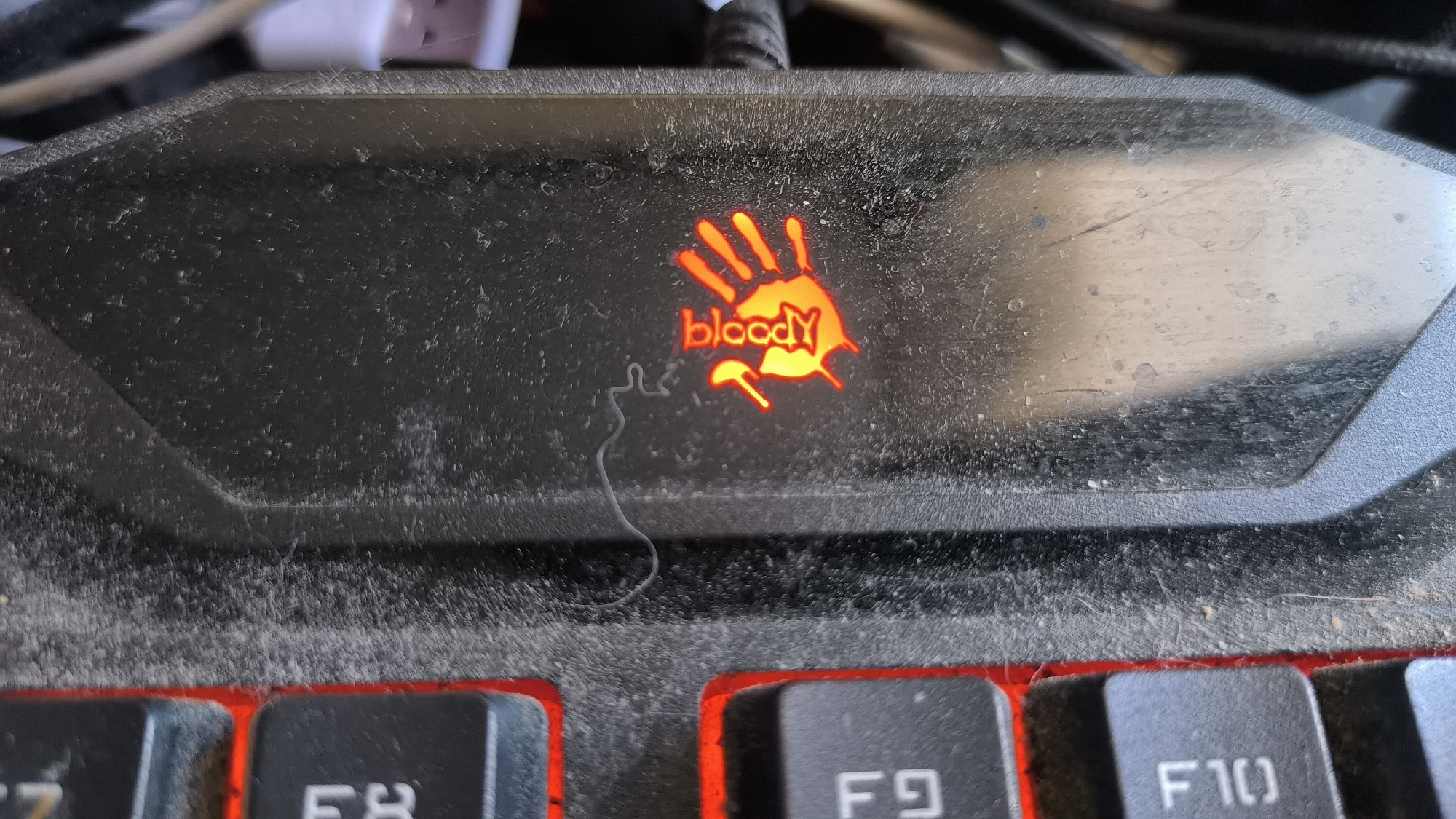This media file is a close-up, eye-level photograph of a dirty, black gaming keyboard, likely taken indoors. The primary subject is the upper portion of the keyboard, specifically the area above the function keys and the branding logo.
The logo, illuminated in orange, features a stylized handprint with dripping lines, accompanied by the word "bloody" written in a matching font below it. The keyboard surface is heavily coated in dust and debris, with visible specks and a long, thin strand of what appears to be hair or thread lying across the logo area. The keys in the foreground, such as F7, F8, F9, and F10, are partially visible, their black surfaces also dusted. The red backlighting of the keys is visible around their edges, contrasting with the dark keyboard body.
In the background, cables and what might be a USB connector are blurred, suggesting the presence of connected peripherals. The lighting appears to be artificial, with no indication of natural light sources. The overall impression is one of a used, perhaps neglected, piece of electronic equipment.
There is no visible text other than the brand name "bloody" on the logo. No people are present, and no specific event or activity is depicted beyond the static image of the keyboard. The location is specified as Kovrov, Russia, but there are no visual cues in the image to confirm this. The time of day and weather are not discernible from the image content.
