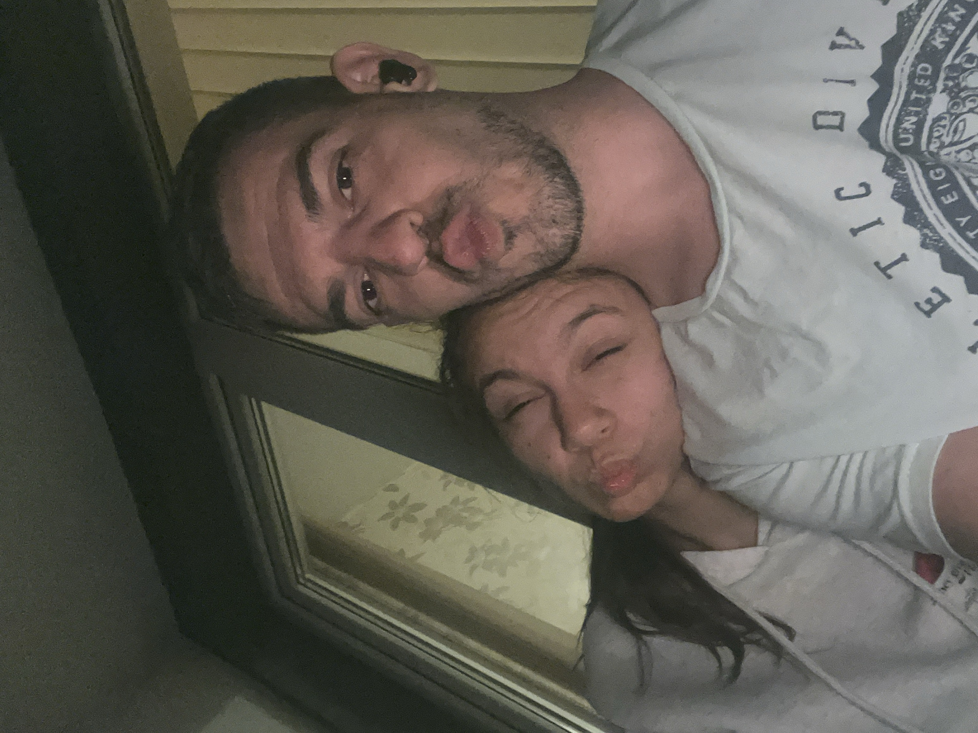 In a close-up selfie taken indoors, likely in an apartment in Sofia, Bulgaria, a man and a woman are depicted. The man, facing forward with his mouth puckered, is positioned in the foreground. He has short, dark hair, stubble, and is wearing a light blue t-shirt with text. An earbud is visible in his right ear. The woman is leaning her head on his shoulder, her eyes closed and her mouth also puckered. She has long, dark hair and is wearing a light-colored hoodie. The background shows a window with blinds and a floral wallpaper pattern. The lighting suggests it is either evening or nighttime.