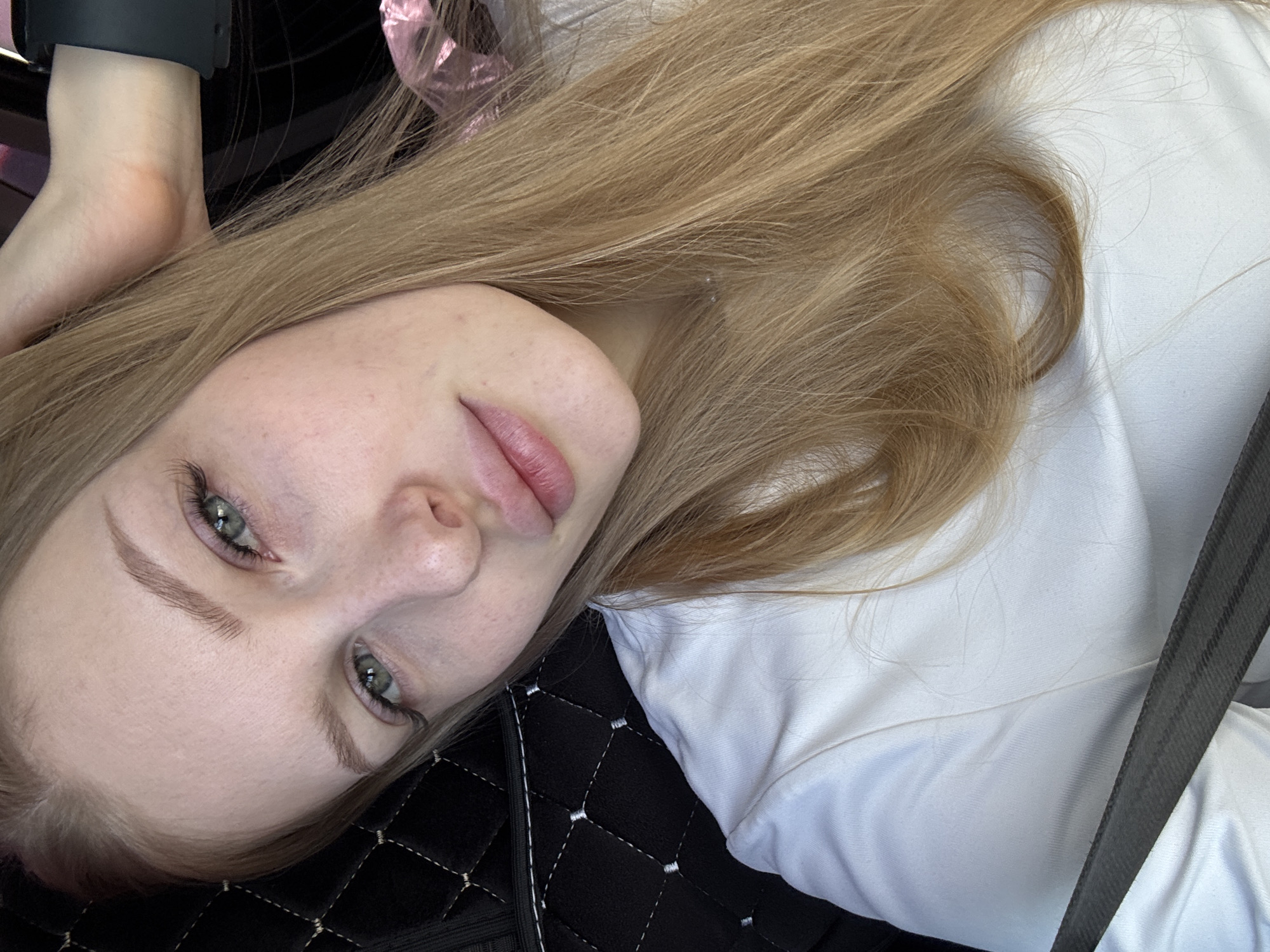 A close-up, angled portrait features a young woman inside what appears to be a car in Perm, Russia. She has long, light brown or blonde hair and striking greenish-grey eyes, which are directed towards the viewer with a calm, neutral expression. She wears a plain white top, and a dark grey or black seatbelt diagonally crosses her chest.
Her skin is fair and appears clear, with subtle makeup highlighting her eyes and lips. Her eyebrows are well-defined. In the upper left corner, part of her left arm is visible, adorned with a black wristband or device. A slight orange tint is noticeable on her skin near the wrist. Behind her arm, a soft pink, somewhat reflective material, possibly part of a decoration or packaging, is partially visible. The background consists of a dark, quilted surface, likely the interior of the vehicle, with white stitching creating a diamond pattern. No visible text is present in the image. The lighting is soft and diffused, suggesting an indoor setting during the daytime.
