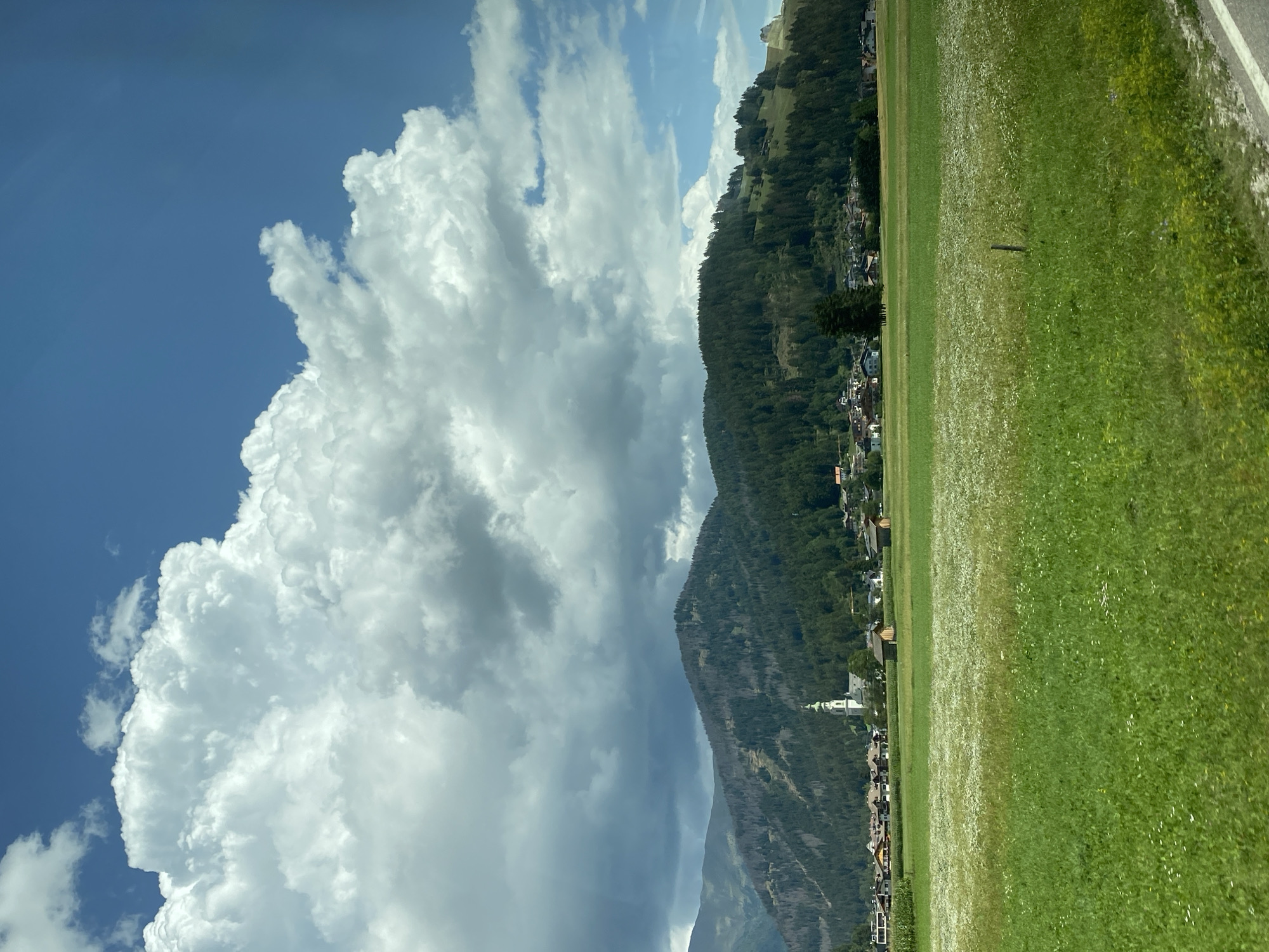 The image depicts a panoramic view of the outskirts of Toblach (Dobbiaco), Italy, under a dynamic sky. In the foreground, a lush green meadow is dotted with numerous small white wildflowers, stretching towards a line of scattered buildings. Beyond the meadow, a village is nestled at the base of a verdant, tree-covered mountain range. A prominent white church steeple with a green roof rises above the village houses. The mountain slopes show some signs of damage, with areas of sparser tree cover. Dark, dramatic storm clouds gather in the upper portion of the sky, contrasting with patches of bright blue sky. The overall scene captures a rural landscape with signs of impending weather.