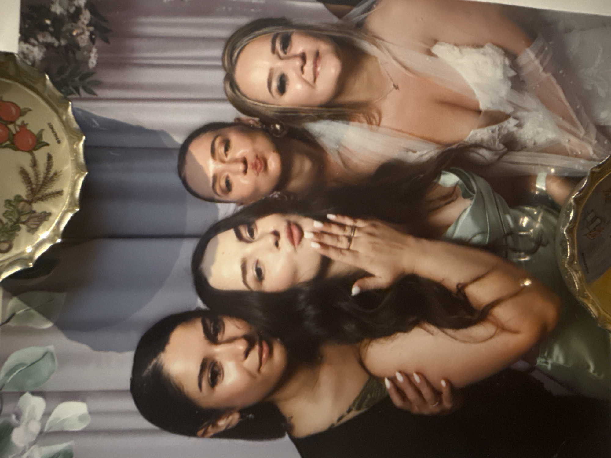 Four women are posing for a photo booth picture. The woman on the far left is wearing a dark v-neck top with a green floral pattern underneath. The woman next to her is wearing a light green strapless dress and blowing a kiss. The third woman from the left is wearing a sheer white veil and making a kissing face. The woman on the far right is wearing a white lace dress with a veil and looking at the camera. The background appears to be a curtain with floral accents. The image is a bit blurry and has a vintage filter applied. The location is not specified in the image or its metadata.