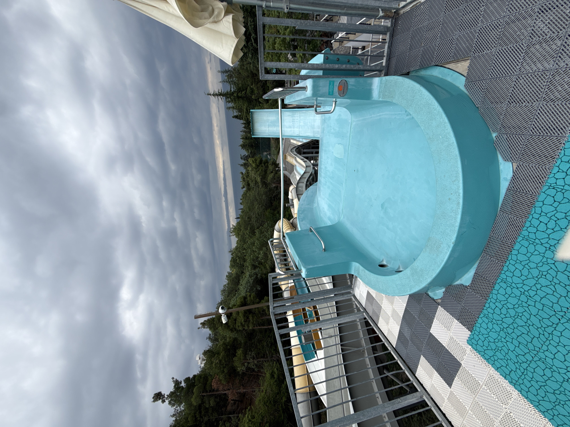 The image shows the top of a blue water slide at a water park. The slide is a bright blue, circular basin with a slightly raised edge. Beyond the slide, there are other water slides, some of which appear to be enclosed tubes. The water park is situated on a hillside covered in lush green trees. In the distance, a body of water, possibly the Black Sea, can be seen under a cloudy, overcast sky. The overall impression is of a slightly damp and moody day at an outdoor attraction. The specific location in Yalta, Ukraine is not definitively identifiable from the visual information provided in the image.