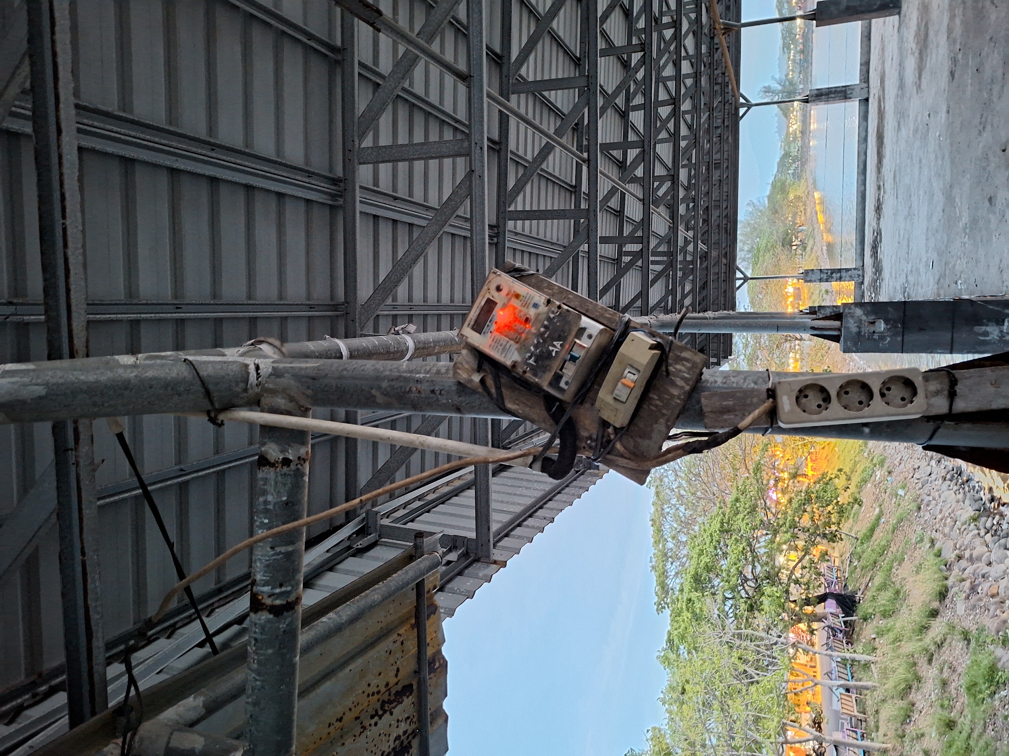 The image shows an outdoor electrical setup attached to a metal pipe support structure. The setup includes a fuse box and a triple power outlet, both secured to the pipe with tape and wiring. The electrical components appear weathered and possibly aged.
The structure is located outdoors, near a paved area and a landscaped section with trees and grass. The backdrop includes a body of water and distant buildings or structures, suggesting a location within a city.
The scene is in Lhokseumawe, Indonesia. The lighting indicates it is likely daytime. The image lacks any visible people or specific actions.