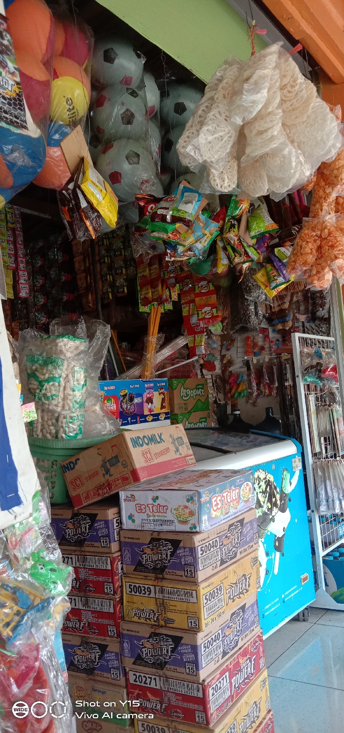 A small store displays various goods. Hanging from the ceiling are deflated soccer balls, snacks, and puffed rice. Below, a refrigerator covered with an ice cream advertisement is stacked with boxes of Power F energy drink and Indomilk Strawberry milk. Jars of snacks are arranged on top. The image includes product prices and the camera information "Shot on Y15 Vivo Al camera."