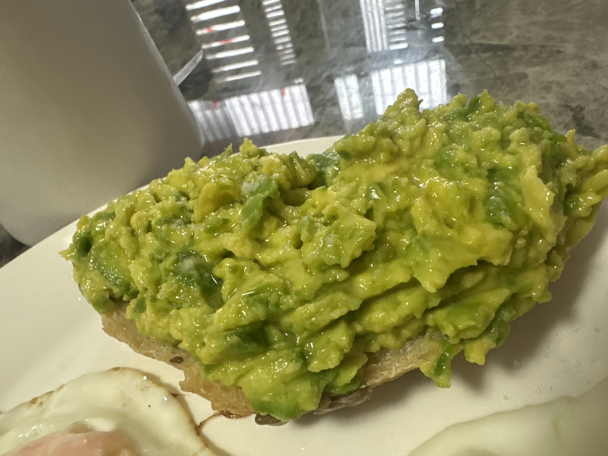 A close-up shows a slice of toast heavily topped with mashed avocado. Part of a fried egg is visible on the left side of the plate. A white mug sits out of focus on the left. The background includes reflections and a view through blinds or shutters. The scene appears to be indoors. The image does not definitively show the location in Bac Nha Trang Ward, Vietnam.