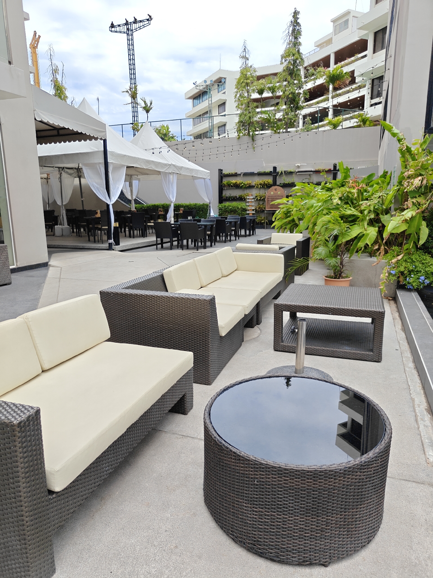 An outdoor patio dining and lounge area is depicted in Pattaya City, Thailand. The scene includes several white tents with decorative draping set up over tables and chairs, suggesting a covered outdoor dining space. In the foreground, a large, dark wicker sectional sofa with light-colored cushions faces a dark wicker coffee table with a reflective glass top. To the right, a dark wicker rectangular coffee table with a lower shelf stands near lush green plants in the background. The area is paved with light-colored concrete. In the distance, a tall lighting tower with multiple lights is visible, along with multi-story buildings that appear to be hotels or condominiums. The sky is cloudy. There are no people visible in the image.