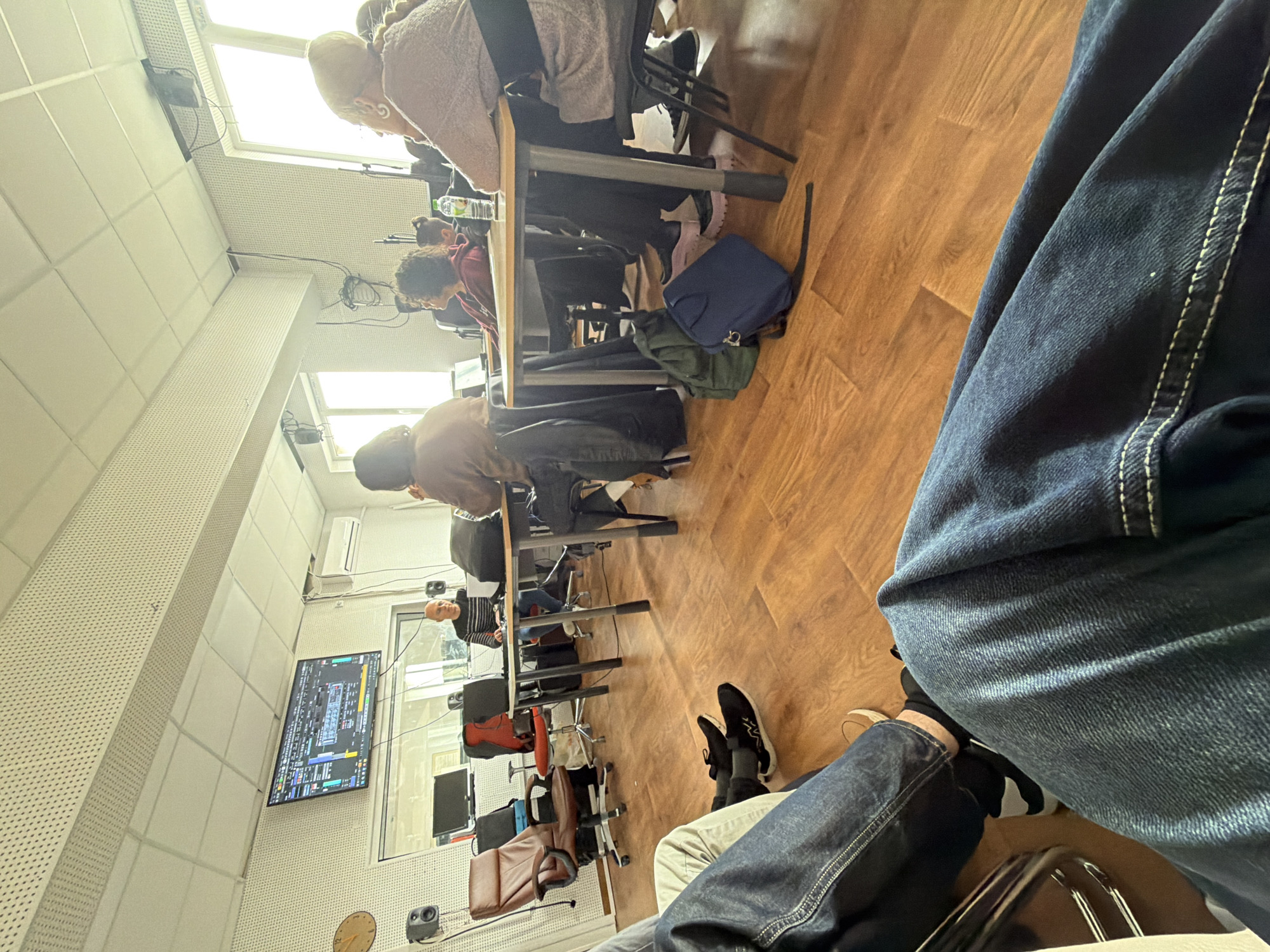 The image shows an indoor scene in what appears to be a classroom or a training facility. Several people are seated at desks, seemingly engaged in some form of learning or work activity. A large display screen is visible on one of the walls, showing technical data or a simulation. Chairs, desks, and personal items like bags and jackets are scattered throughout the room. The flooring is wooden. The image is taken from the perspective of someone sitting in a chair, with their legs and part of their clothing visible in the foreground. I cannot ascertain the location in Sofia, Bulgaria, from the visual information provided.