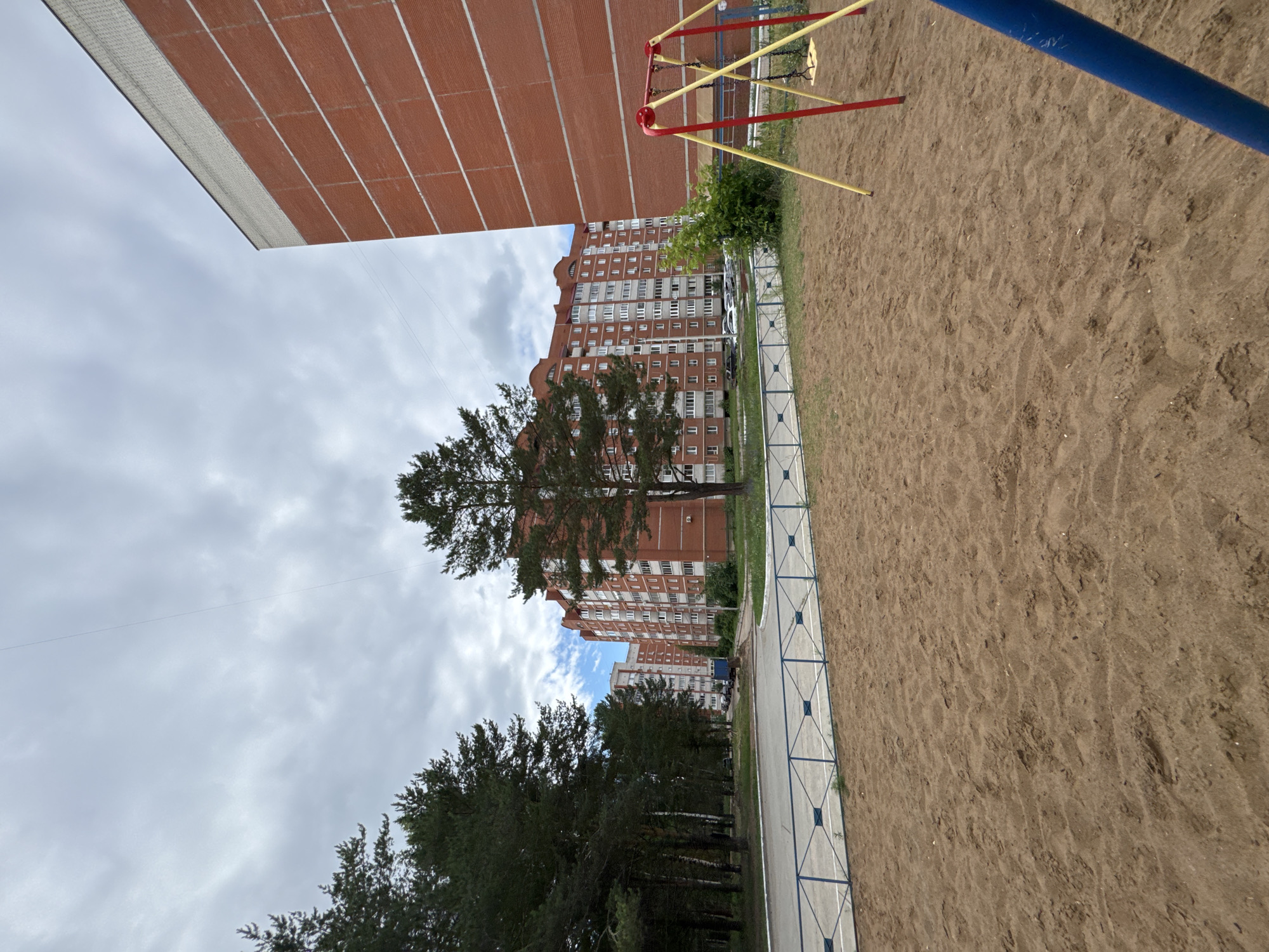 The image depicts a public outdoor space in Chaikovsky, Russia, featuring a playground area in the foreground and residential buildings in the background. The playground has a sand-covered ground, a blue and yellow swing set with a black seat, and a blue pole from another play structure partially visible on the right. A light blue fence with diamond-shaped openings separates the playground from a paved area with a road. In the mid-ground, a row of tall, multi-story brick apartment buildings with white-framed windows and balconies stretches across the scene. Mature pine trees line the left side of the image, with a few smaller trees and bushes interspersed throughout the area. The sky is overcast with scattered clouds. The overall impression is one of a residential neighborhood with recreational facilities.