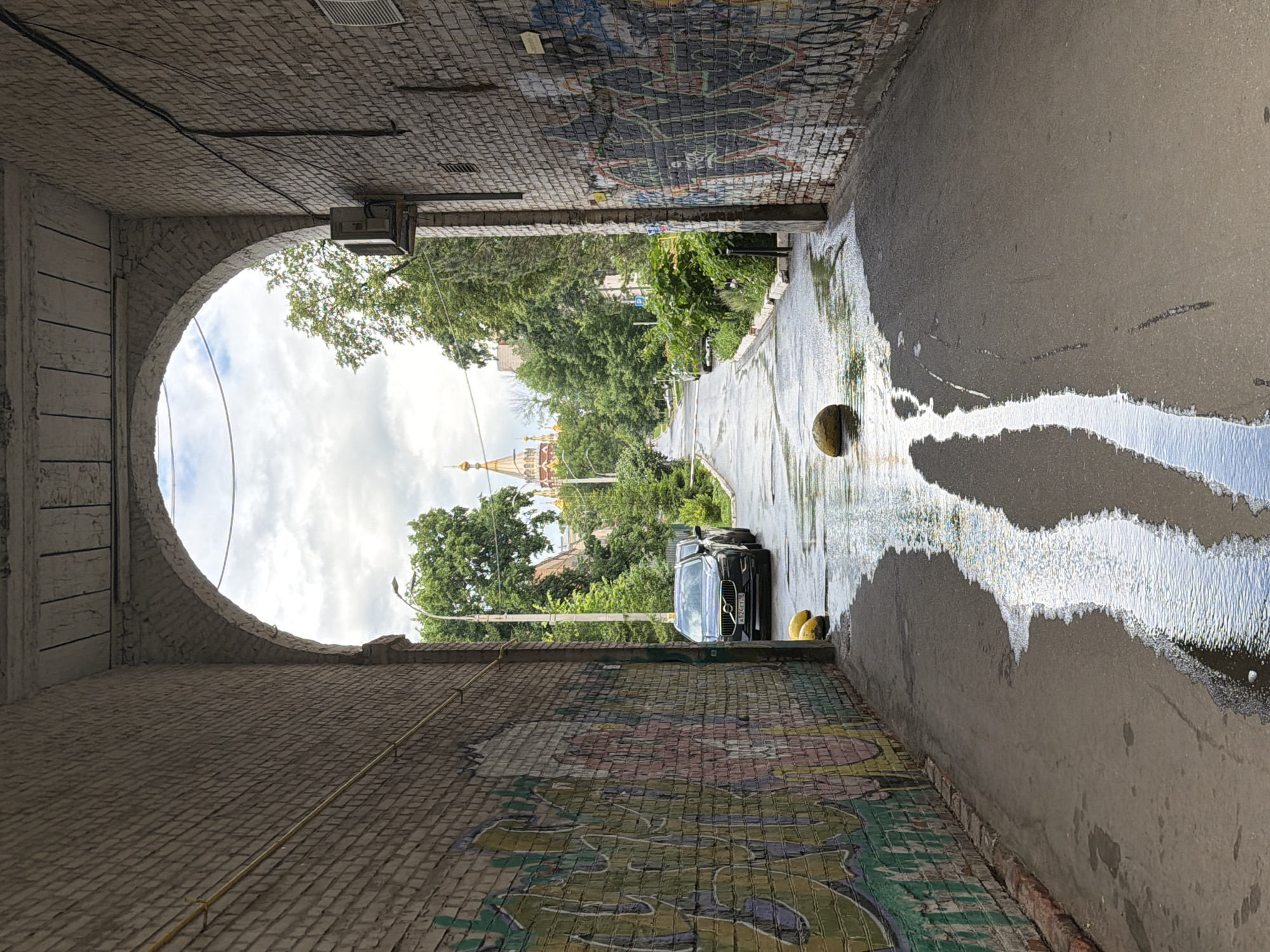 This is a photograph taken from within an archway leading out onto a street in Izhevsk, Russia. The archway is constructed of brick and shows signs of wear and tear, with peeling paint on the ceiling. The walls of the archway are covered in graffiti.
Outside the archway, the street is wet, suggesting recent rain, and there are puddles on the asphalt. A dark-colored Volvo SUV is parked on the right side of the street, with its license plate visible. Yellow bollards are positioned along the side of the road. In the distance, beyond the trees lining the street, a church with golden domes is visible. The sky is overcast.
The photograph is taken from a low angle, looking forward through the archway. The lighting suggests it is daytime. There are no people visible in the frame.