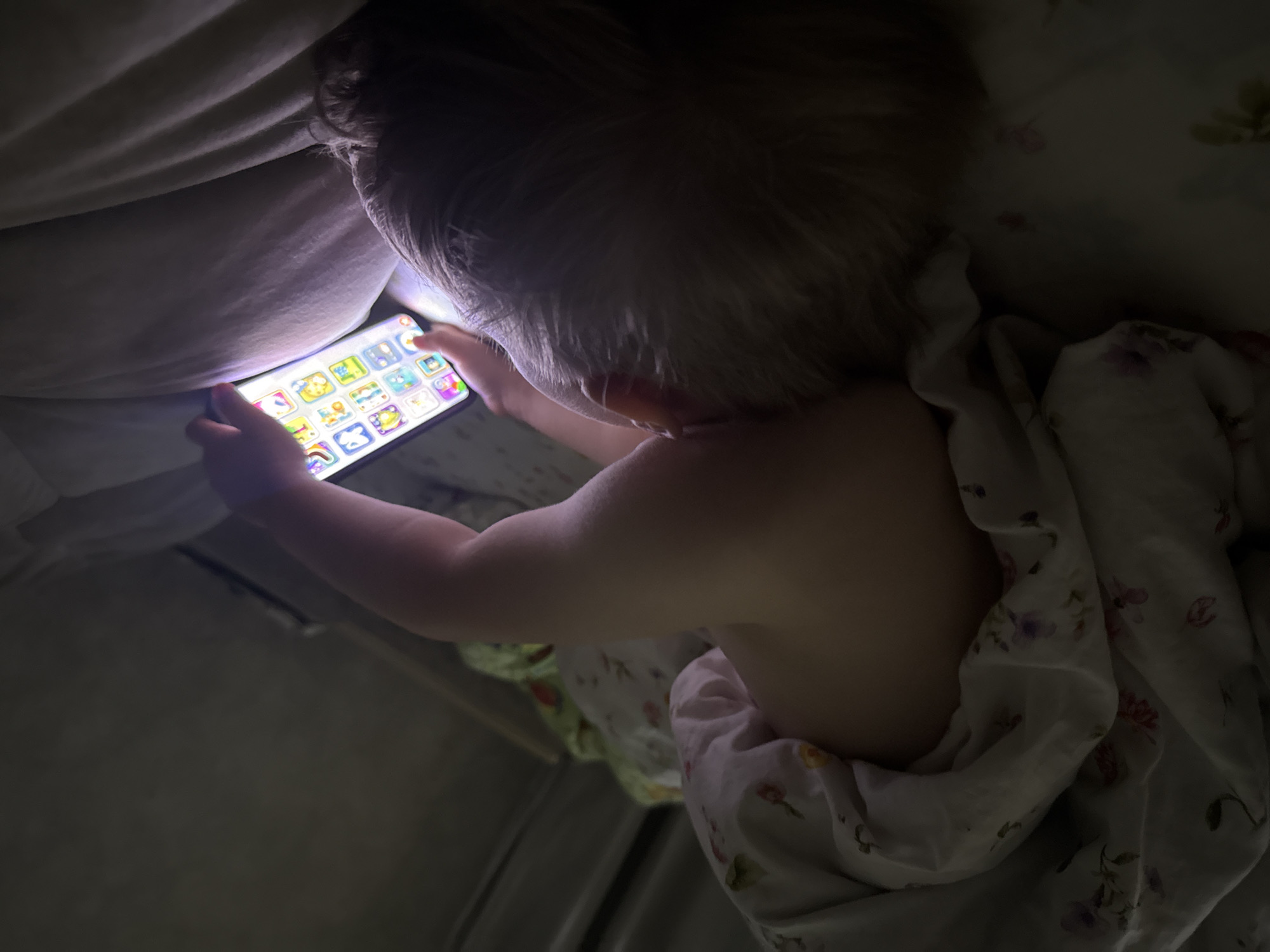 A child with blond hair is lying on their back in bed, holding a smartphone. The phone screen is bright and displays multiple colorful icons. The child's right arm is extended, and their fingers are touching the screen. The child is partially covered by a floral-patterned blanket. The lighting in the room is dim, with light emanating from the phone screen and a light source above and to the right of the child. The background is dark and appears to be a wall or headboard. This scene is not identifiable as being in Октябрьское, Russia.