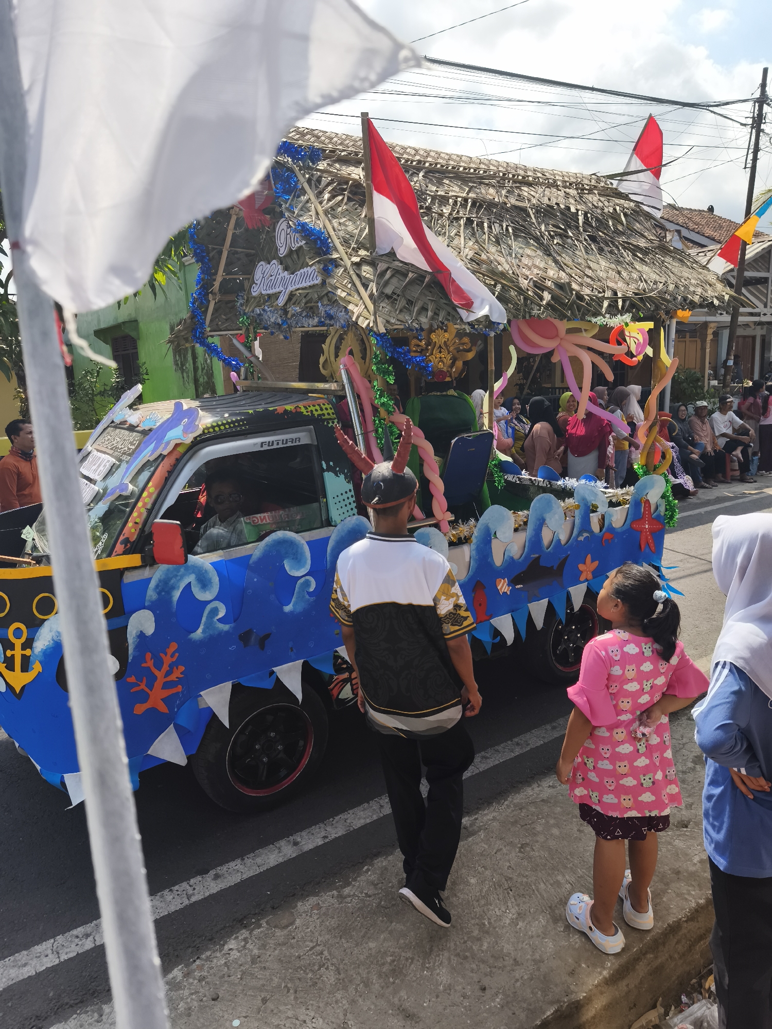 In Kutoarjo, Indonesia, a vibrant parade float, adorned with a traditional thatched roof and decorated with Indonesian flags and blue tinsel, moves down a street. The float is mounted on a pickup truck, its sides painted with a blue wave design featuring marine life like a dolphin, shark, fish, and starfish, as well as an anchor. A driver wearing sunglasses is visible inside the truck's cabin. Several people, including a man in a black and white patterned shirt and black pants, and a young girl in a pink owl-patterned dress, stand on the roadside observing the parade. Spectators are also seated on benches along the street. The scene captures a lively community event under a bright, partly cloudy sky.