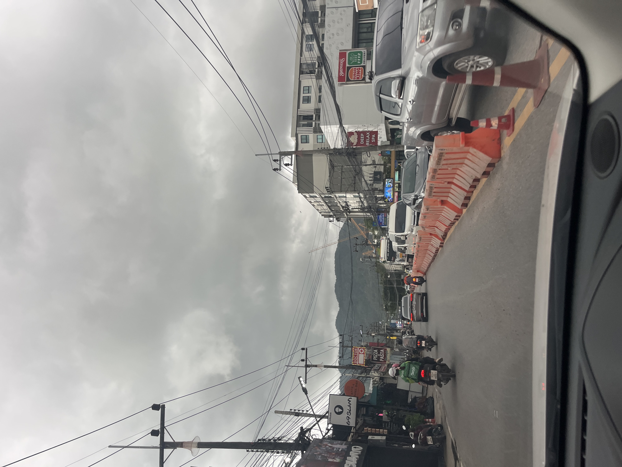 The image depicts a street scene in Choeng Thale, Thailand, under an overcast sky. The perspective is from inside a vehicle, looking forward. The road is lined with orange construction barriers on the right side, separating traffic lanes. Several vehicles are present, including a silver pickup truck, cars, and motorcycles. On the left side of the road are buildings with various signs, including one for "69SLAM" and another with "PIZZA PASTA." A person wearing a green Grab delivery uniform is riding a motorcycle. In the background, there are buildings and mountains, with some construction cranes visible. The overall atmosphere suggests a moderately busy street with ongoing infrastructure work.