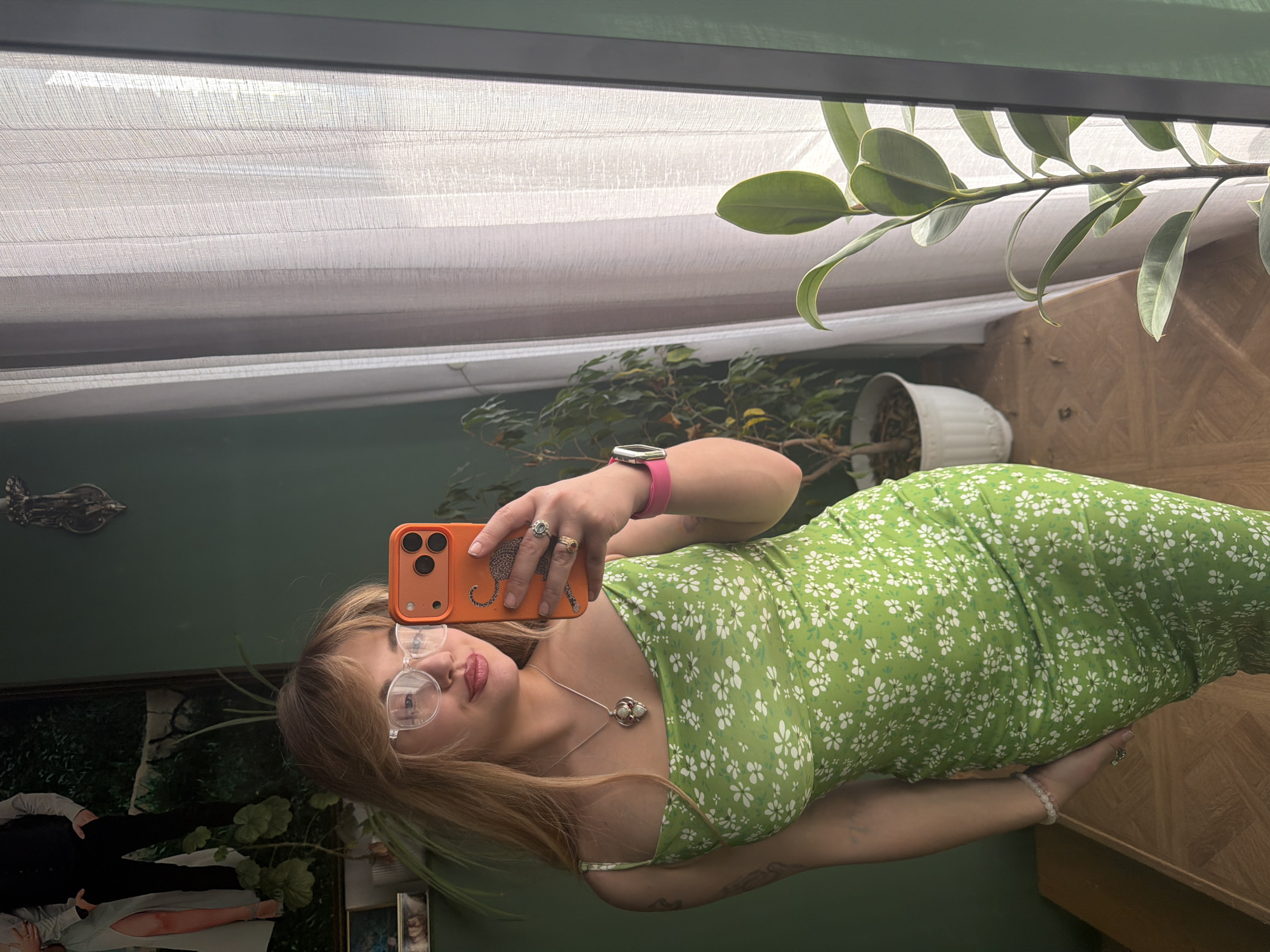 A woman takes a selfie in a mirror indoors. She is wearing round glasses, a green dress with white floral patterns, and a watch. Her phone has an orange case. The reflection shows various decorative items, plants, and a painting. The setting seems to be a room in a house, possibly in Большая Шильна, Russia, given the location data.