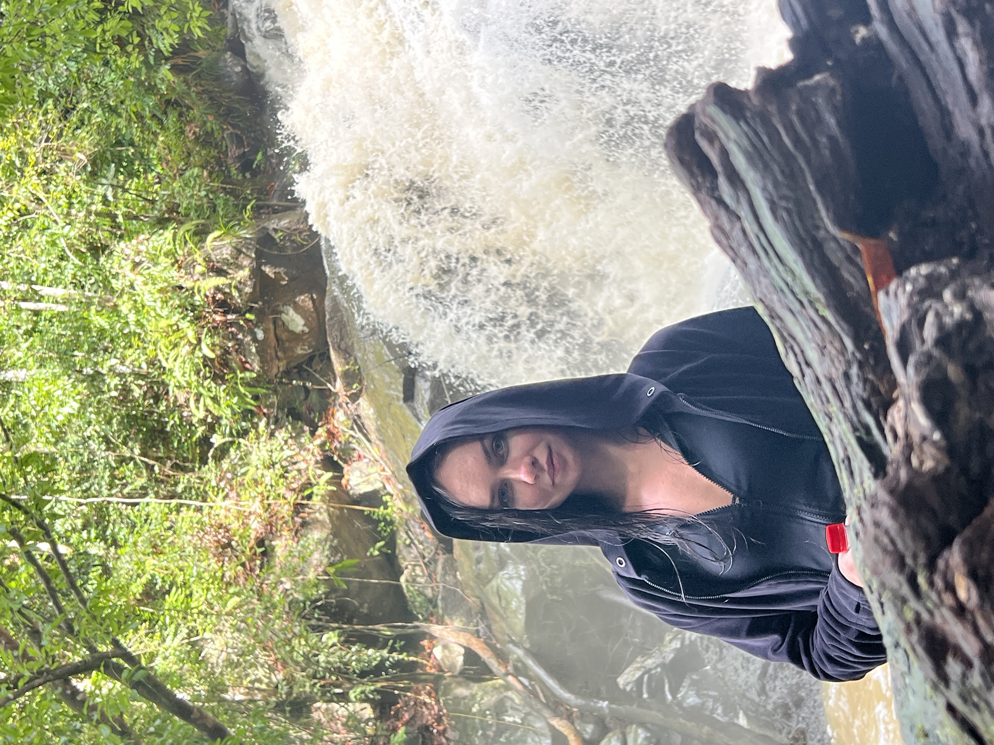 A young woman is captured in a portrait-style photograph, posing in front of a powerful waterfall in a lush, natural setting in Siyambalagoda, Sri Lanka. She appears to be in her 20s or 30s, positioned on the right side of the frame, looking directly at the camera with a neutral and calm expression. She is wearing a dark, possibly black or navy blue, hooded jacket with the hood up, and her dark hair appears damp, likely from the waterfall's spray.
A prominent, dark-toned tree trunk or rock formation occupies the immediate foreground on the right, partially obscuring the woman's arm. In her right hand, partially visible behind this foreground element, she holds a small red object, which might be a bottle cap or a small container.
Behind the woman and extending across the left side of the frame, a large and energetic waterfall cascades downwards, creating a turbulent expanse of white, foamy water. The air around the falls appears misty. To the far left and behind the waterfall, dense, vibrant green vegetation, including trees and bushes, thrives, indicating a rich, forested environment. Rocks and water are visible at the base of the falls. The scene is illuminated by natural daylight, suggesting it is daytime. There is no visible text in the image. The photograph conveys a sense of serene engagement with nature amidst a striking natural landmark.