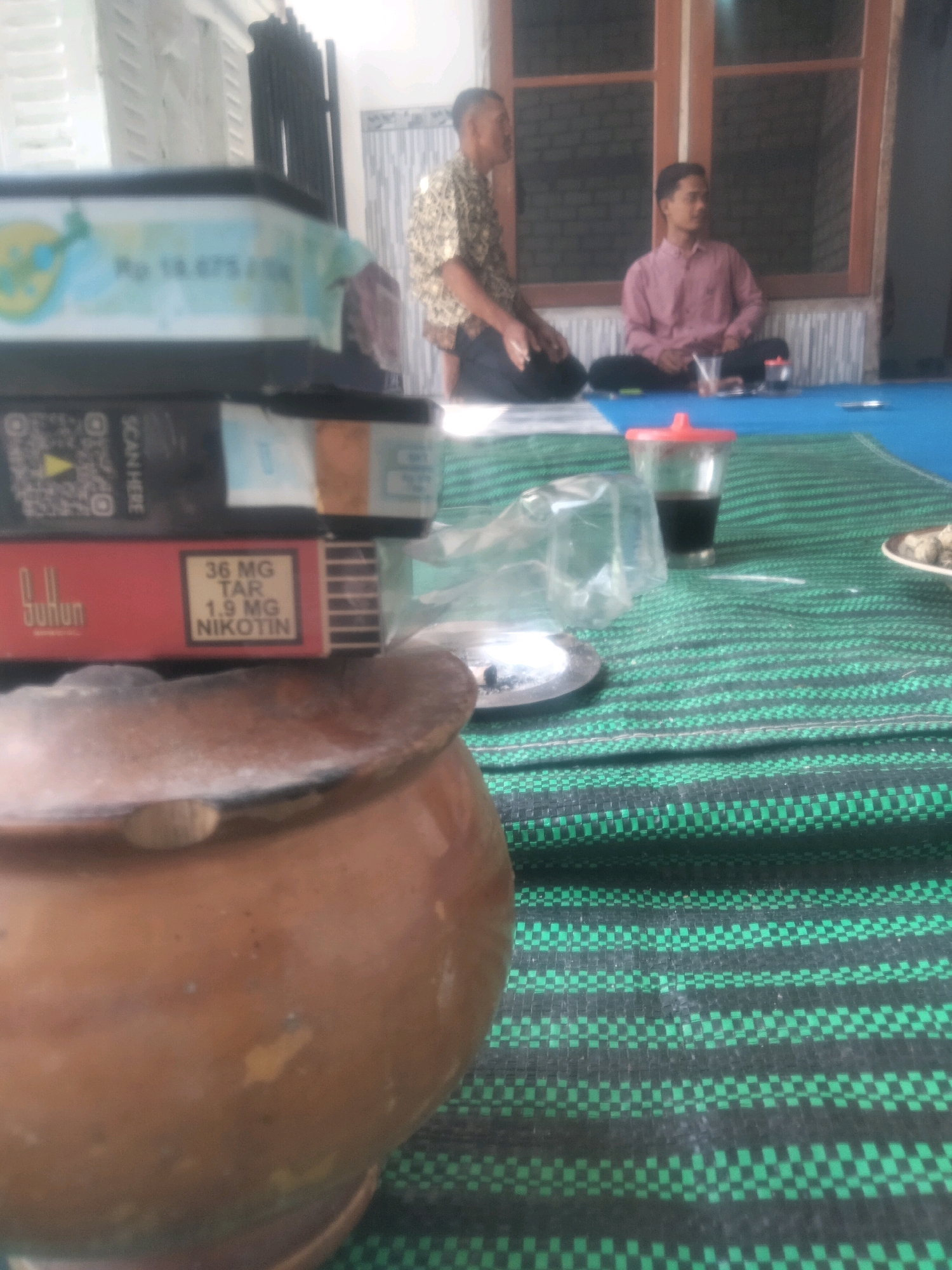 In Soko, Indonesia, an outdoor scene unfolds with two men seated on the floor. The man on the left wears a patterned short-sleeved shirt and dark pants, while the man on the right sports a pink long-sleeved shirt and dark pants. They are positioned on a blue mat that is partially covered by a green and black woven mat. In the foreground, a brown clay pot is prominently displayed. To the right of the pot, on the woven mat, are several items including cigarette packs, a plastic bag, an ashtray, a glass of dark liquid with a red lid, and a small plate with food. The background features a brick wall with a wooden-framed window, and a white wall with horizontal siding. The lighting suggests it is daytime.