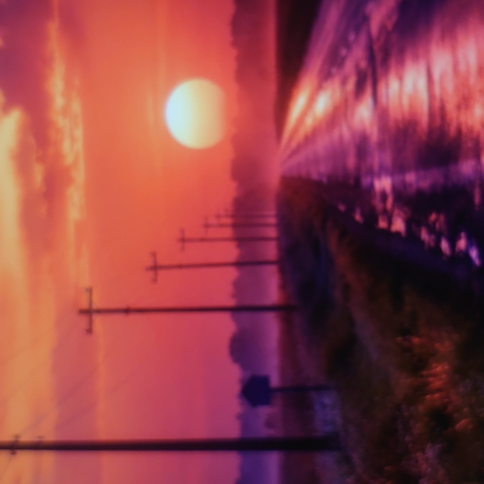 The image depicts a sunset scene with a road stretching into the distance. The sky is a vibrant mix of orange and pink hues, with a large, luminous sun partially visible. Several telephone poles line the left side of the road, receding into the distance. The road itself appears wet, reflecting the colors of the sky. In the foreground on the left, there is a dark silhouette of what appears to be a mailbox or small structure on a pole. The environment appears rural or suburban, with a line of trees or foliage visible in the background. The overall impression is one of a tranquil, atmospheric sunset. There is no identifiable location in the city of Belorechensk, Russia, visible in the image.