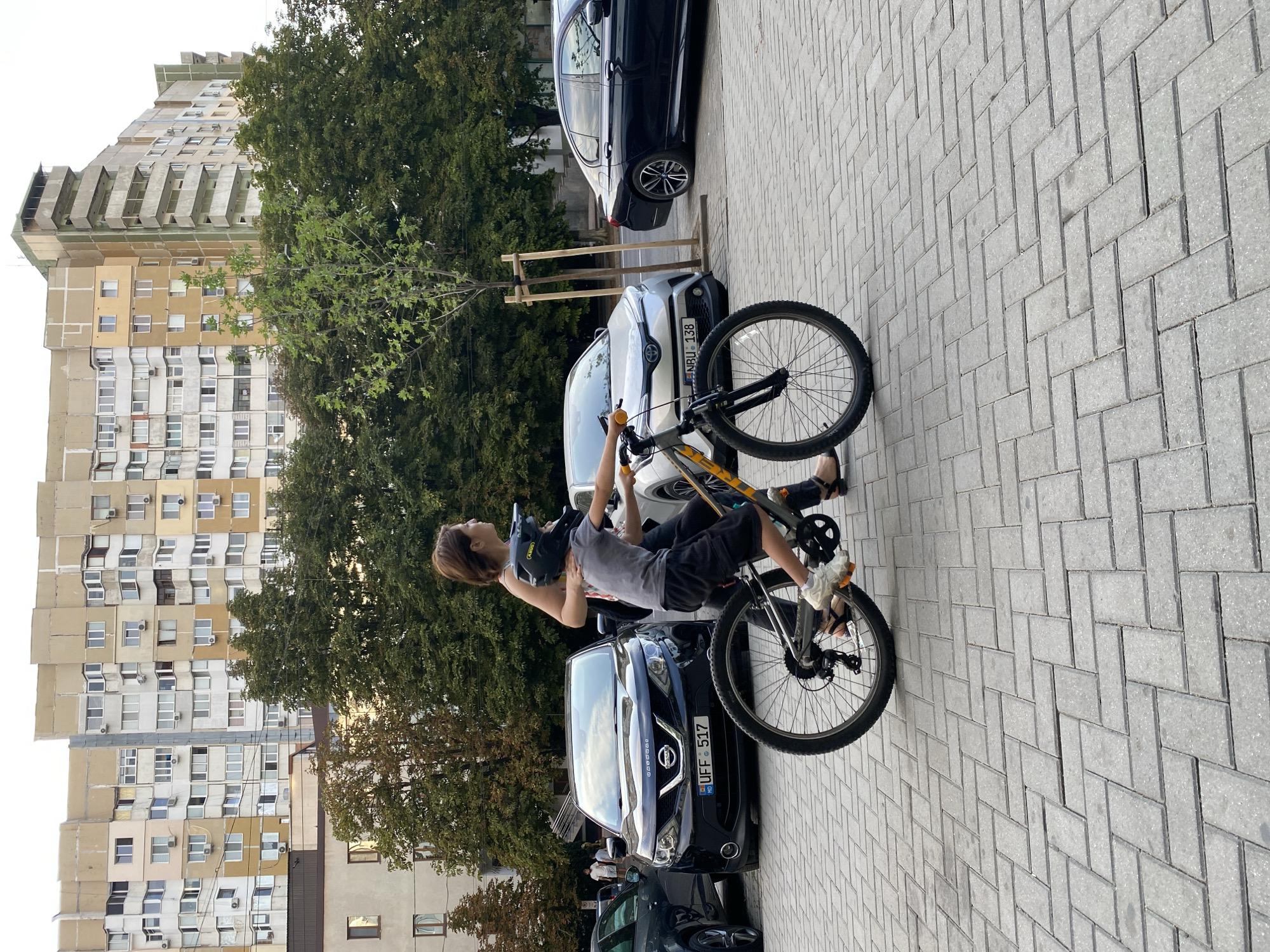 A woman is assisting a young boy on a bicycle on a paved sidewalk in front of parked cars and residential buildings. The woman is wearing a sleeveless top and dark pants, and the boy is wearing a grey t-shirt, dark shorts, a helmet, and white shoes. The bicycle is grey and orange. In the background, there are several cars, including a black Nissan with a Moldovan license plate, a silver car, and a black sedan. Behind the cars and trees, there are multi-story apartment buildings. The sidewalk is made of grey interlocking pavers. The scene appears to be in Chișinău, Moldova, given the visible license plate of the black Nissan.
