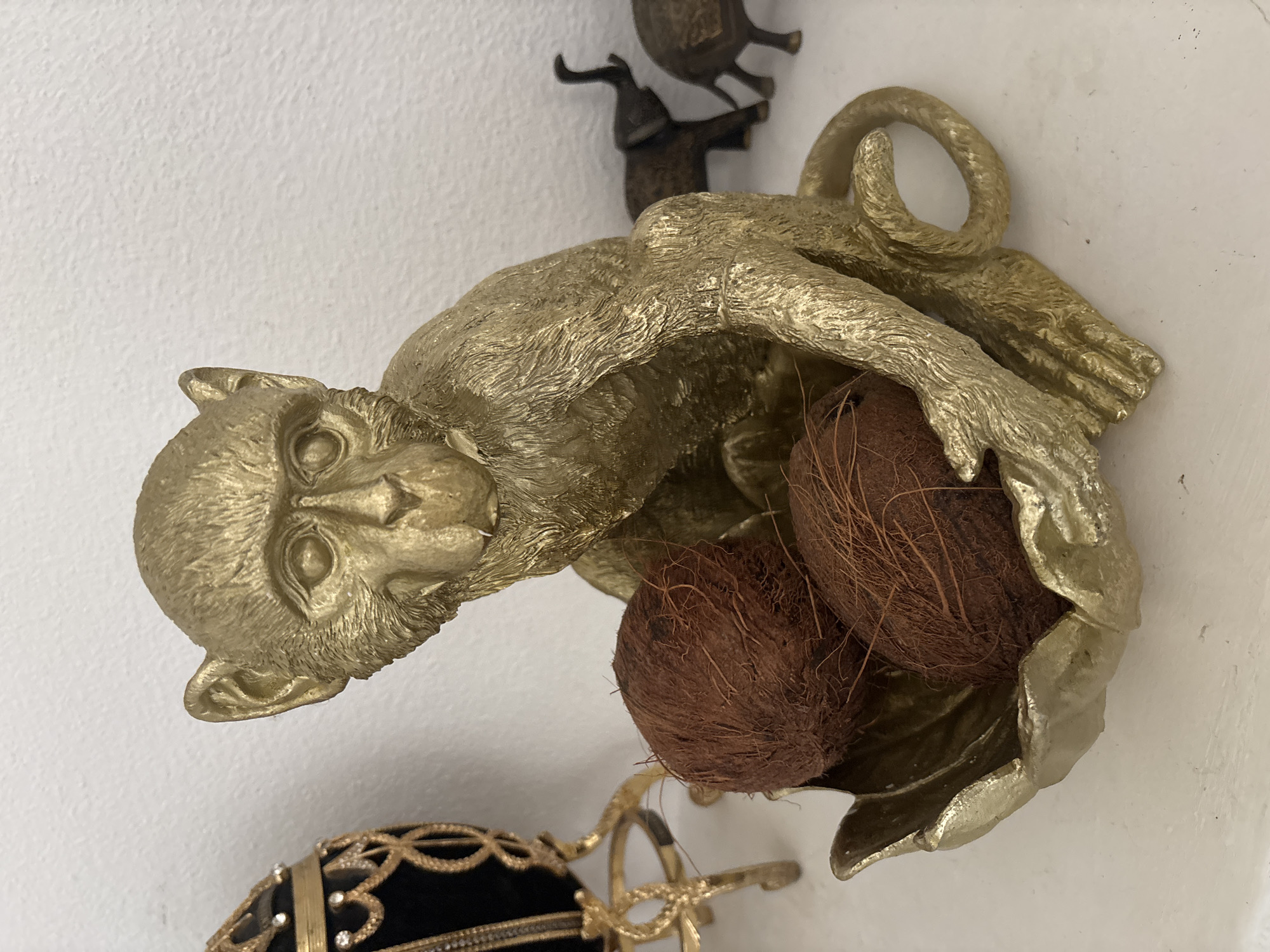 A gold-colored monkey figurine sits on a white surface, cradling three coconuts in its hands. The monkey is facing forward with its head tilted slightly. Behind the monkey, to the left, is a decorative black and gold egg on a stand. To the right, a small herd of dark-colored elephant figurines are visible. The background is a textured white wall. The scene appears to be an indoor display, possibly in a home or shop. The specific location within the city of Orihuela, Spain, cannot be determined from the image alone.