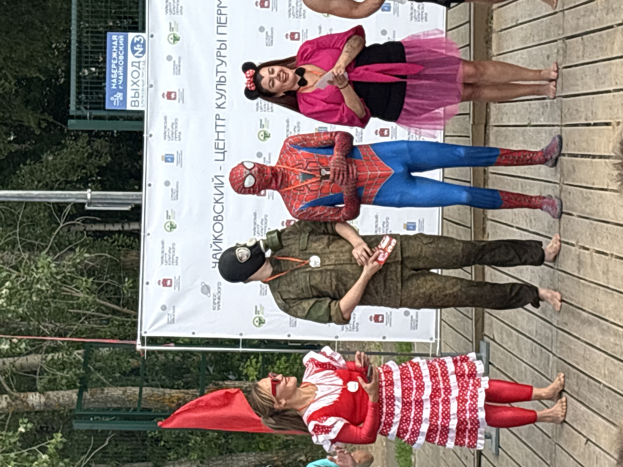 The image shows a group of people in costumes standing on a wooden platform in front of a large banner. From top to bottom, the individuals are dressed as Minnie Mouse, Spider-Man, a soldier with a gas mask, and a person in a red and white striped dress. The Minnie Mouse character, a woman, is smiling and holding papers. Spider-Man is standing with his arms crossed. The soldier is holding a small box. The person in the striped dress is also smiling and wearing red sunglasses.
The banner behind them has Cyrillic text, including "ЧАЙКОВСКИЙ - ЦЕНТР КУЛЬТУРЫ ПЕРМ" which translates to "Chaikovsky - Center of Culture Perm." A blue sign with "НАБЕРЕЖНАЯ Г.ЧАЙКОВСКИЙ ВЫХОД №3" (Chaikovsky Embankment Exit No. 3) is visible to the left. The scene appears to be an outdoor event, possibly a festival or a gathering, with trees and foliage visible in the background. The lighting suggests it is daytime, and the weather seems to be pleasant. The overall mood seems festive and engaging.