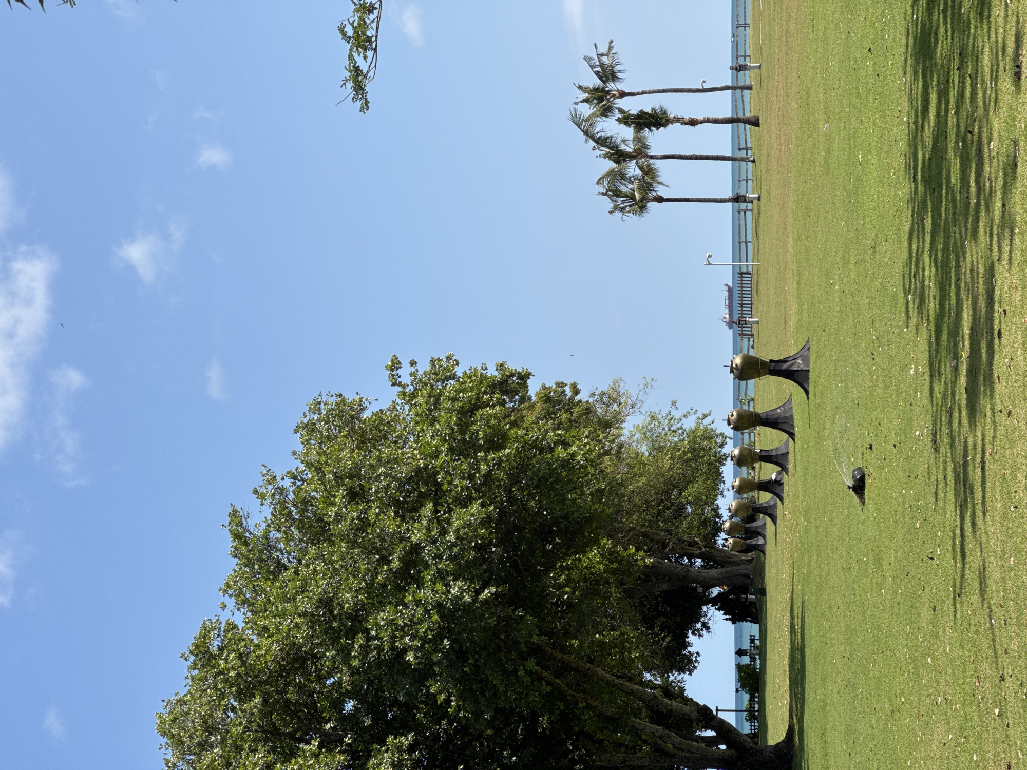 The image shows a grassy area in Kuta Selatan, Indonesia, likely a park or public space. A row of distinctive metallic sculptures, resembling stylized horns or speakers, lines the edge of the grass near a row of trees. In the background, there are several palm trees situated near the coastline. A body of water and distant ships are also visible. The sky is blue with scattered clouds.