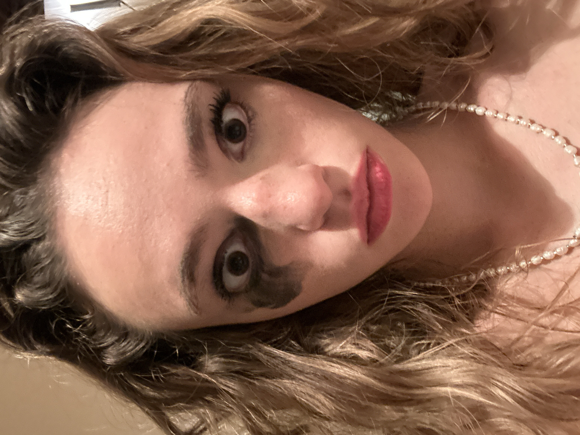 The image is a close-up selfie of a young woman with curly, blonde hair. She is wearing a pearl necklace. Her makeup is smudged around her eyes, giving her a disheveled appearance. Her eyes are wide open, and her mouth is slightly open in a pout. The background is a plain, light-colored wall. There is no indication of the location being San Bernardino Tlaxcalancingo, Mexico.
