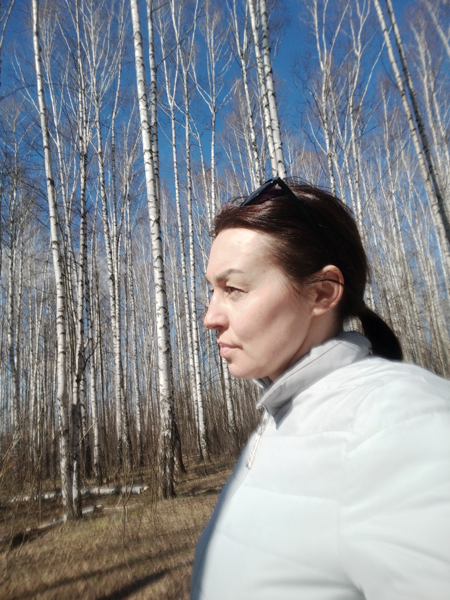 A woman stands in a birch forest in Chaikovsky, Russia. She has brown hair pulled back in a ponytail and is wearing a white puffer jacket. Sunglasses rest on top of her head. The trees are tall and slender, with white bark and black markings. The ground is covered in brown leaves and dry grass. The sky is blue and mostly clear. The woman is facing to the left, away from the camera, with a serene expression.