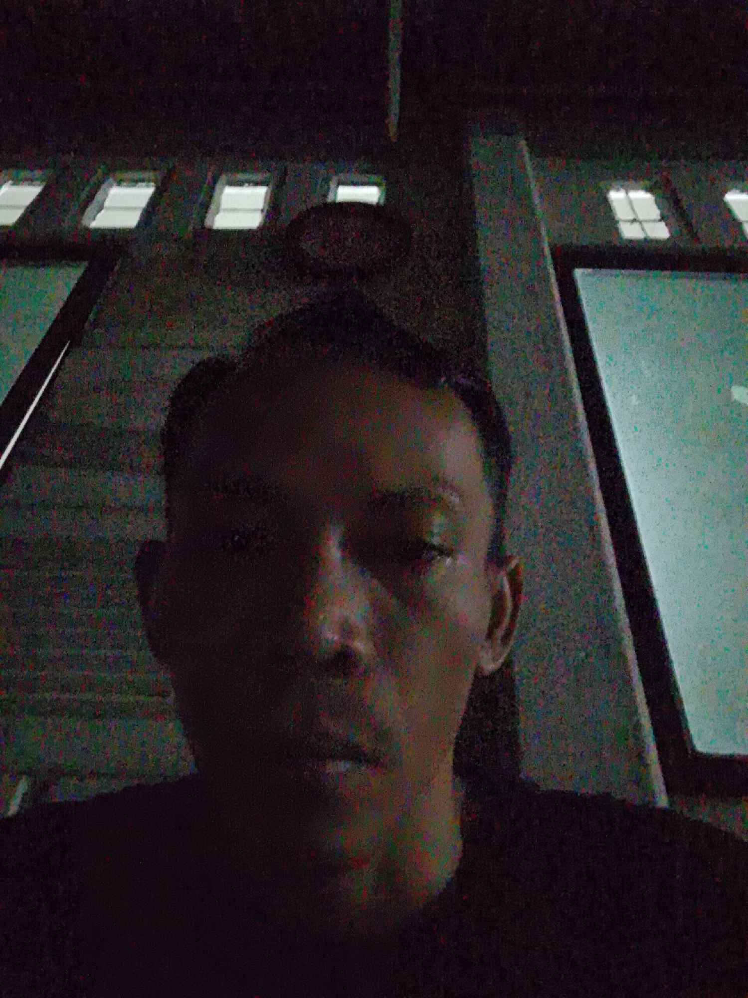 The image depicts a man in front of a building at night, likely in an urban setting. The man is in the foreground, facing the camera, with a dark shirt. The background features a building facade with a series of windows. The location might be Bukit Ketok, Indonesia. The image appears to be of poor quality due to low light and noise.