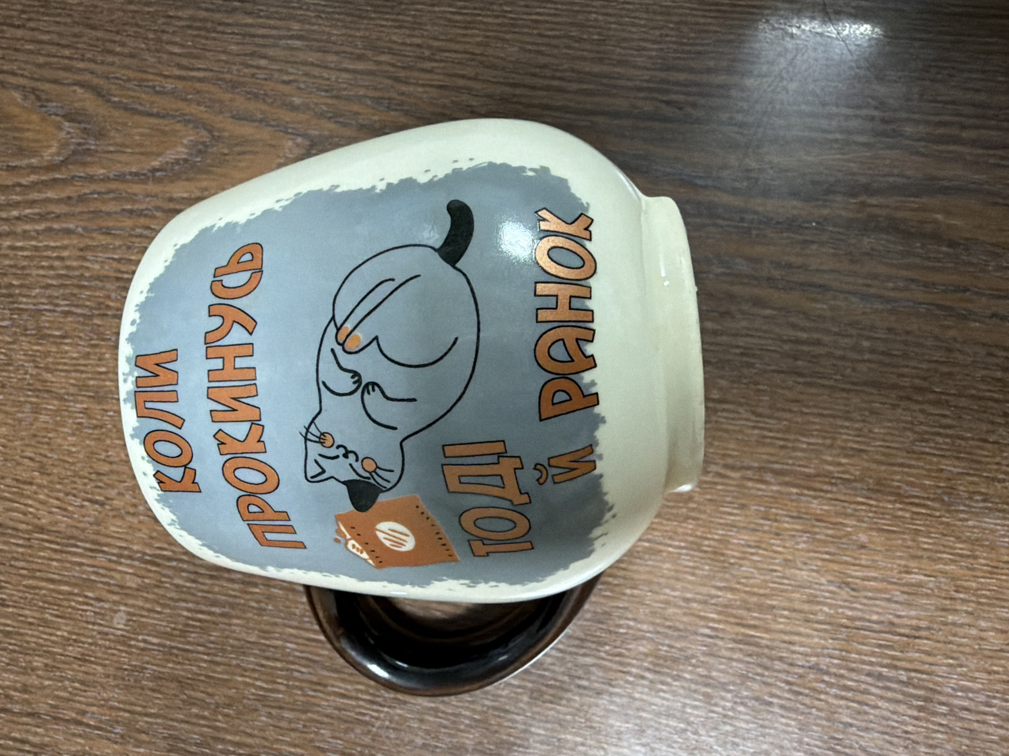 A ceramic mug is depicted lying on its side on a wooden surface. The mug has a creamy white base with a broad, roughly painted light gray band around its middle. On this band, there's a cartoon illustration of a gray cat with a black tail and small orange dots on its cheeks, appearing sleepy or disgruntled as it sits with a small, open, brown book or tablet in front of it.
Flanking the cat illustration is text written in orange-brown Cyrillic letters. On the left, it reads "КОЛИ ПРОКИНУСЬ" (KOLY PROKYNUSʹ), which translates to "When I wake up." On the right, it says "ТОДІ Й РАНОК" (TODI Y RANOK), meaning "Then it's morning." The entire phrase humorously implies that the day only truly begins when the person (or cat) decides to wake up.
The handle of the mug, which appears dark brown or black, is visible underneath the body of the mug. The wooden surface has a visible grain pattern and reflects some ambient light. The overall impression is of a novelty mug with a playful, relatable message for those who struggle with early mornings.