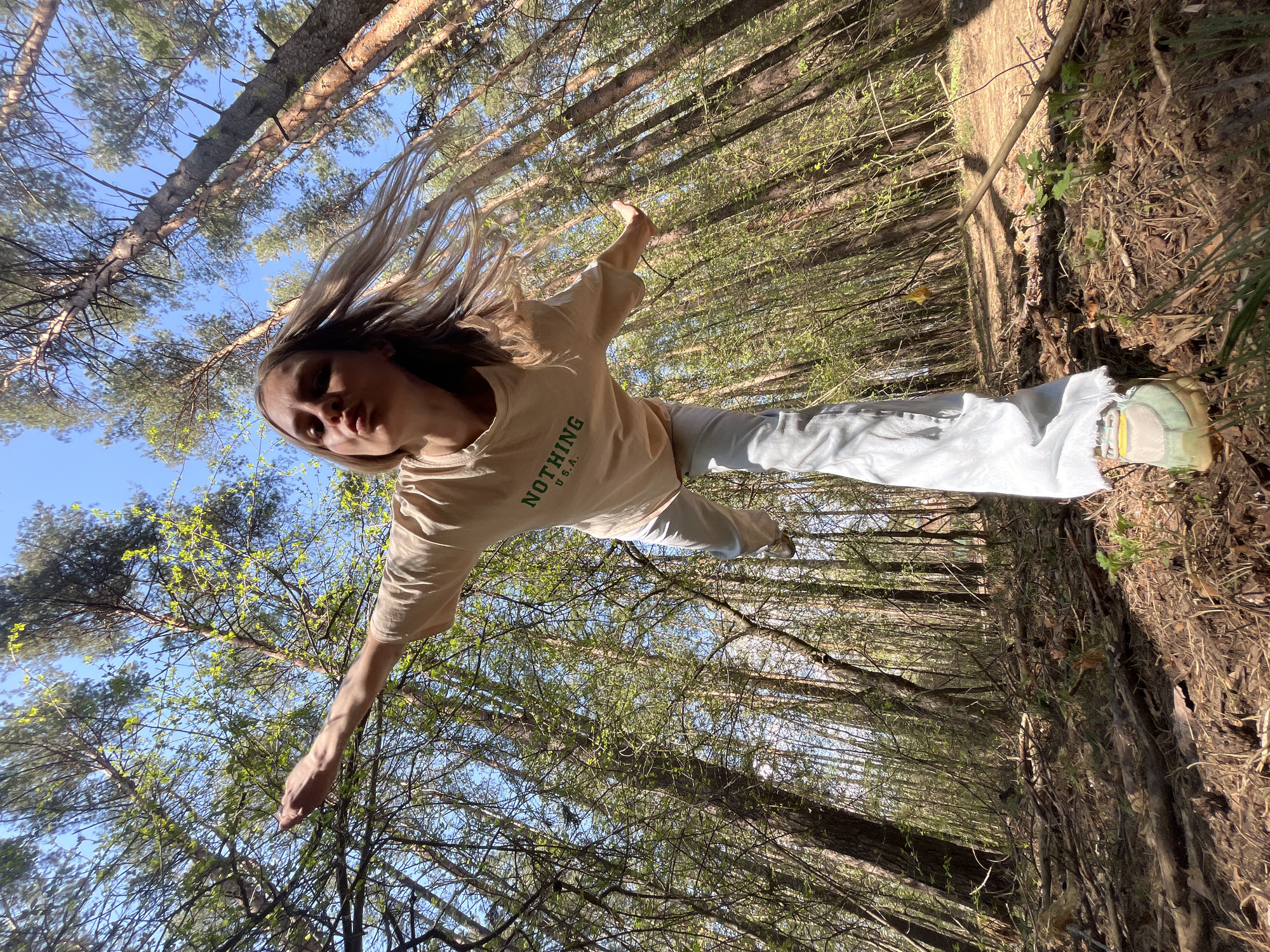 The image shows a woman in a forest. She is wearing a light-colored t-shirt with the word "NOTHING" printed on it and light-colored pants. Her arms are outstretched. The view is from a low angle, looking up through the trees. The trees are tall and thin, with green leaves budding. The sky is visible through the branches. The ground is covered in fallen leaves and dirt.
There is no evidence in the image to determine if this is in the city of Chaikovsky, Russia.