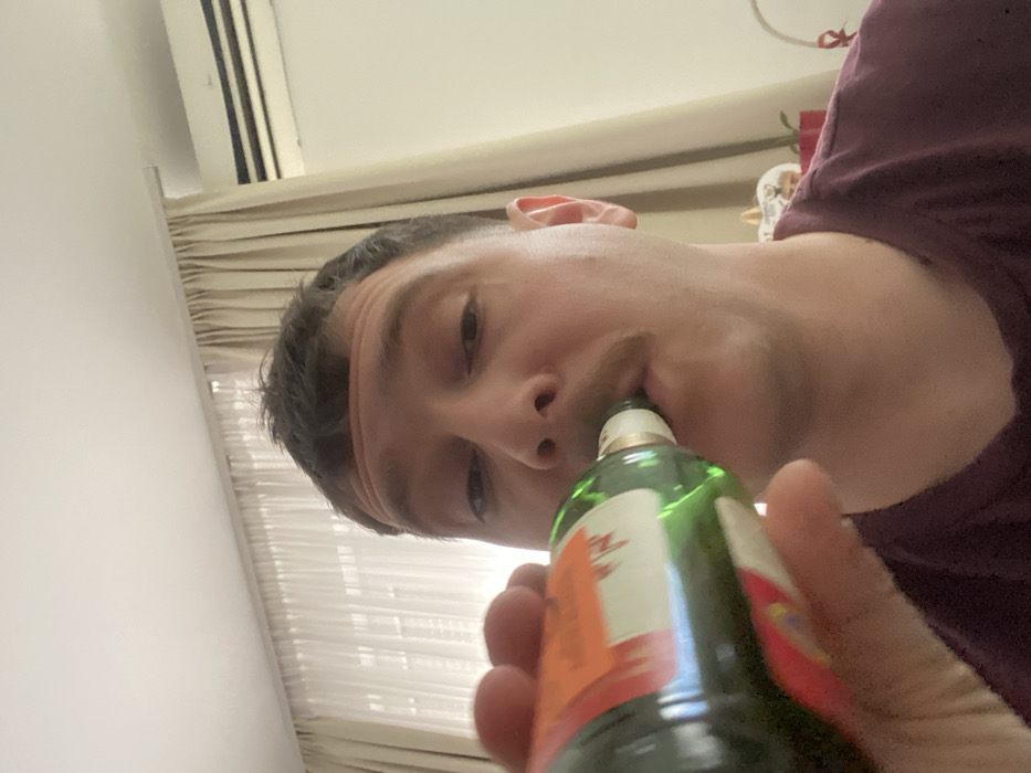 A male individual is depicted in a close-up, taking a drink directly from a green glass bottle within an indoor setting in Vozdovac Urban Municipality, Serbia. The man, with short dark hair and a prominent mustache, is looking directly forward while tilting his head back slightly to drink. He is wearing a dark maroon t-shirt. His right hand grips the mid-section of the bottle. The bottle is a green glass type with an orange, red, and white label, characteristic of Gorki List Pelinkovac. In the background, to the left, light beige, pleated sheer curtains are visible, suggesting a window. To the upper right, a white wall-mounted air conditioning unit is present. A white ceiling is visible above. The lower right portion of the background reveals a blurred white object, possibly a decorative pumpkin, and a red ribbon, suggesting seasonal or decorative elements. The room is brightly lit.