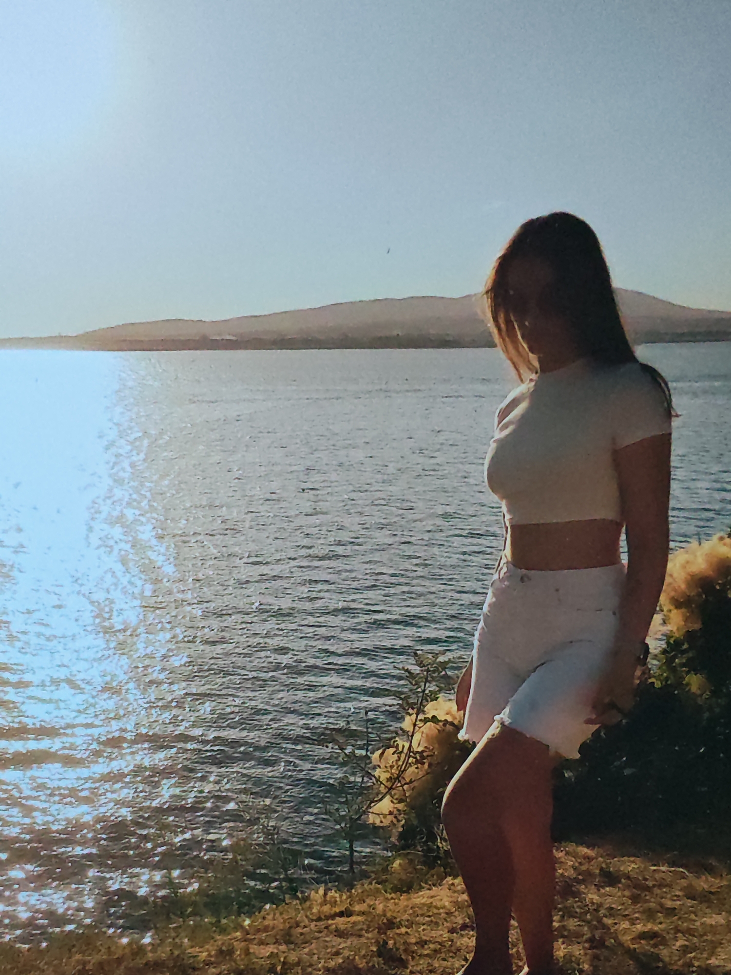 A young woman with long, dark hair stands on a grassy embankment overlooking a wide body of water. She is wearing a white crop top and white denim shorts, with her body angled away from the camera, looking out towards the horizon. The water reflects the bright sunlight, creating a shimmering effect on its surface. In the distance, a range of hills or mountains forms the shoreline. The sky is clear and a pale blue, indicating a sunny day, likely in the late afternoon or early evening given the warm, low angle of the sun casting long shadows and illuminating the water. There are some bushes with yellow flowers and foliage to the right of the woman, adding a touch of color to the scene. The overall atmosphere appears peaceful and serene. No text is visible in the image.
