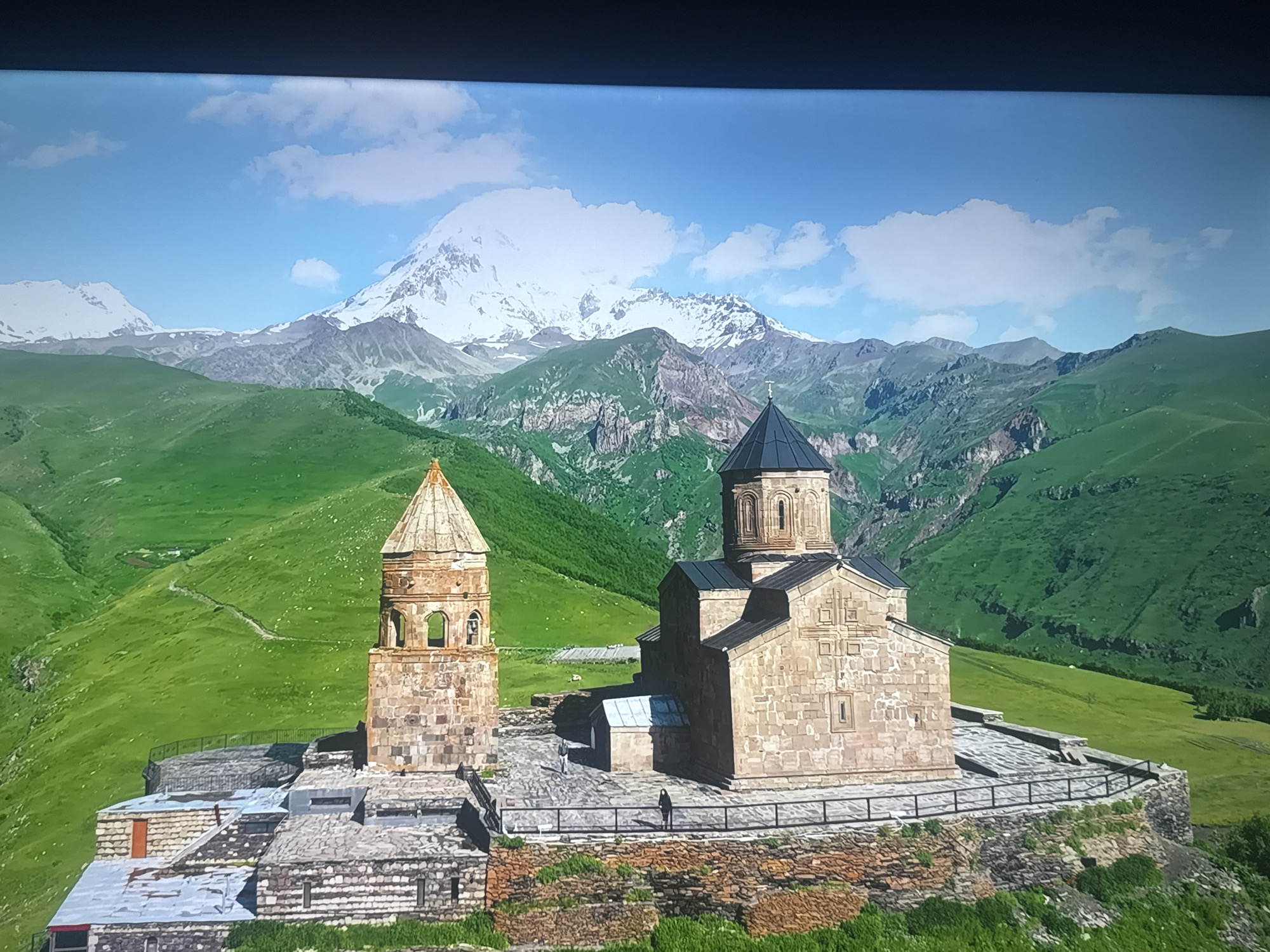 This is a full, eye-level, medium shot of the Gergeti Trinity Church, a 14th-century Orthodox church in Georgia, situated at an altitude of 2,170 meters (7,120 feet) near the town of Stepantsminda. The church is built on a rocky peak, surrounded by lush green mountain slopes. In the background, a snow-capped mountain range dominates the horizon under a bright blue sky with scattered clouds. The church complex consists of a main church building with a dark conical roof and a separate bell tower. Two individuals are visible, one walking near the main church and another standing on a walkway. The scene is bathed in natural light, suggesting a sunny day.