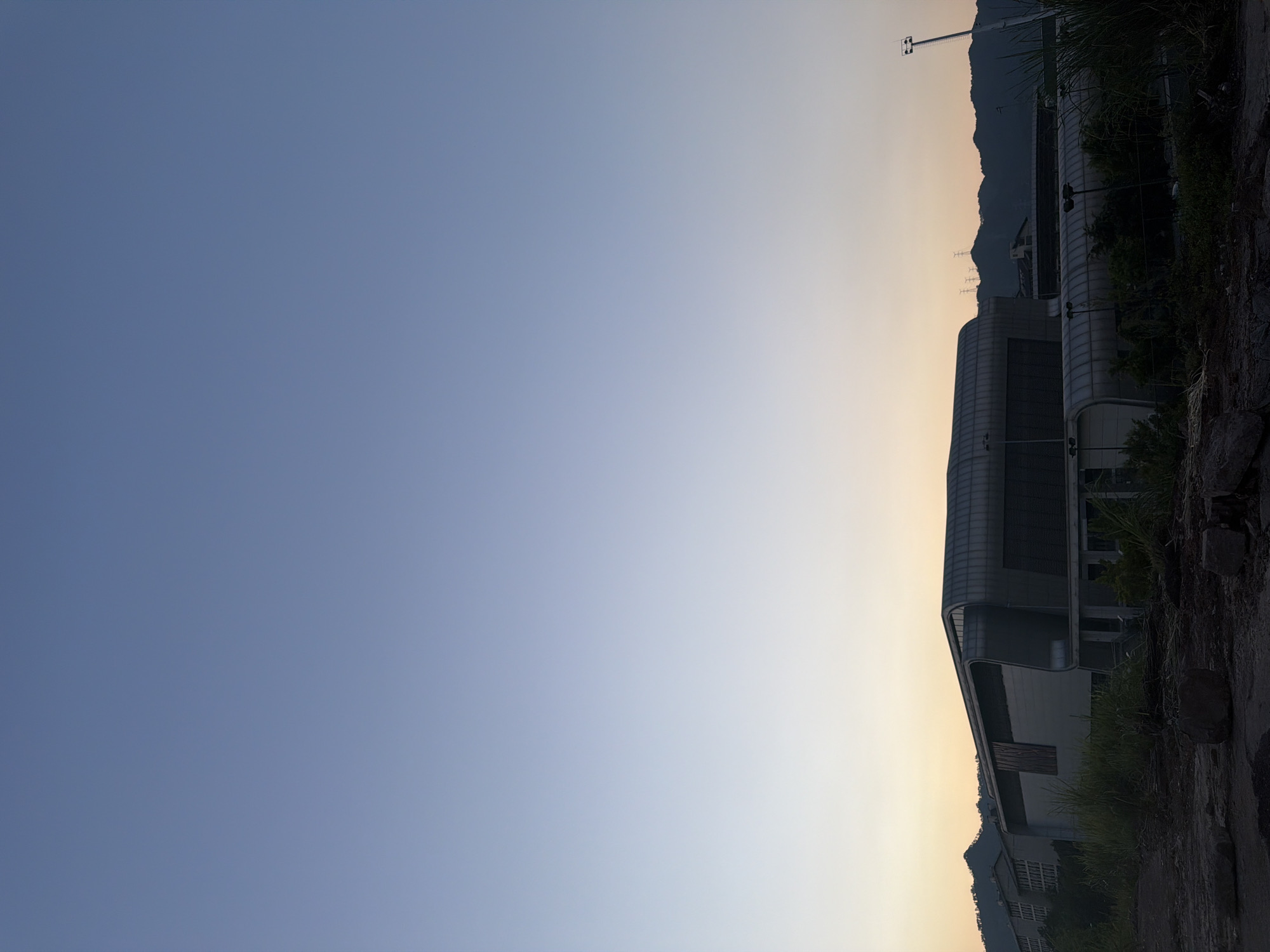 This image depicts an exterior view of a modern, multi-sectioned building with a curved, metallic roof and large glass panels, situated in Jiangjin, China. The building appears to be part of an industrial or commercial complex, with other structures and infrastructure visible in the background, including distant mountains and what seem to be power lines. The foreground shows a rough, unpaved area with scattered rocks and overgrown vegetation, suggesting a partially developed or abandoned site. The sky is clear and features a gradient of pale blue to a soft yellow-orange hue near the horizon, indicating either dawn or dusk. No people are present in the frame. The overall atmosphere is serene and quiet, with a focus on the architectural form of the building against the natural landscape and the atmospheric light.