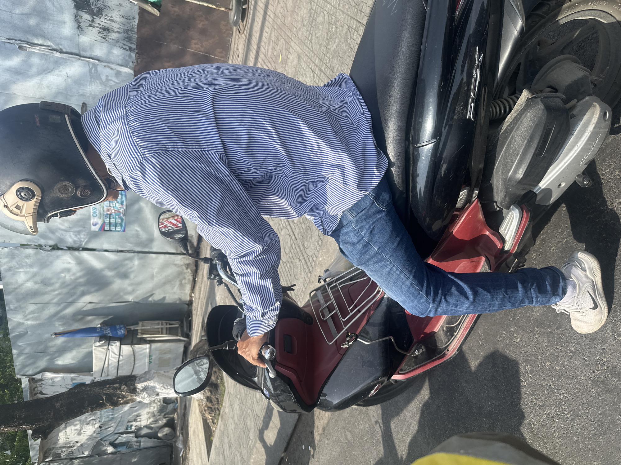A person wearing a blue and white striped long-sleeved shirt, blue jeans, and white Nike sneakers sits on a red and black scooter. The person is wearing a black and beige helmet. The scooter has a chrome rack on the back and the letters "PCX" are visible on the side. The person is holding onto the handlebars of the scooter. The scene appears to be on a street in Bac Nha Trang Ward, Vietnam, with a corrugated metal wall in the background. A tree with peeling bark is to the left, and shadows are cast on the asphalt ground.