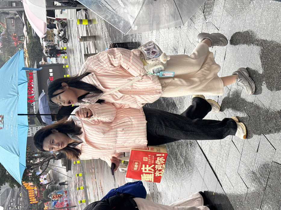 Two people stand in an outdoor environment, likely a street, on what appears to be a wet, overcast day. Both wear pink and white striped jackets. One person wears black pants and white sneakers, holding a red box with Chinese writing. The other wears a cream-colored skirt, a white purse, and gray sneakers. An open blue umbrella is held above them. The background shows a street scene with other people, umbrellas, and various street-side structures.